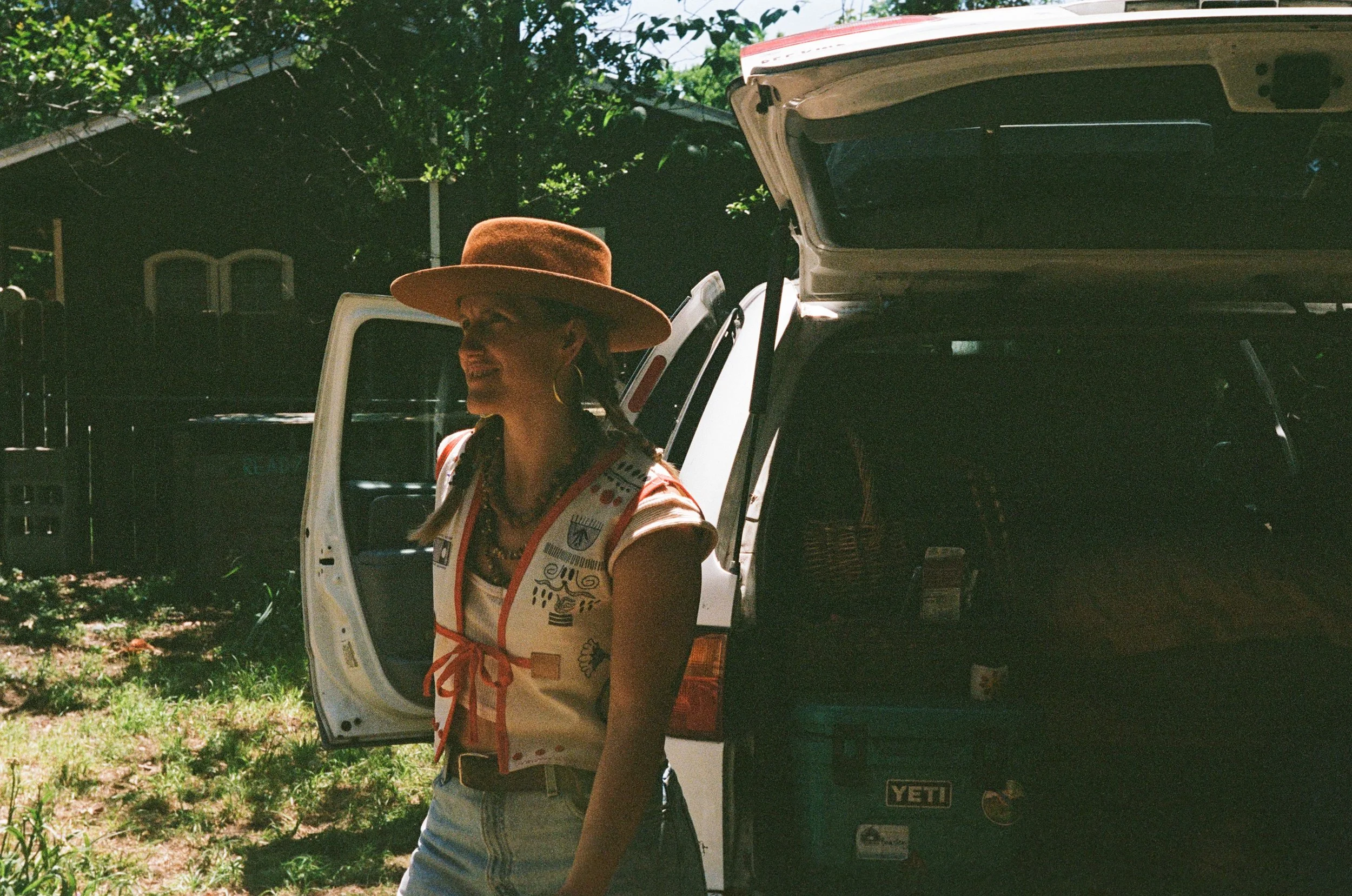 A woman with long braided hair, wearing a large brown hat, sunglasses, hoop earrings, and a colorful vest, smiling while standing outside next to an open vehicle door, with a dark vehicle behind her and green trees in the background.