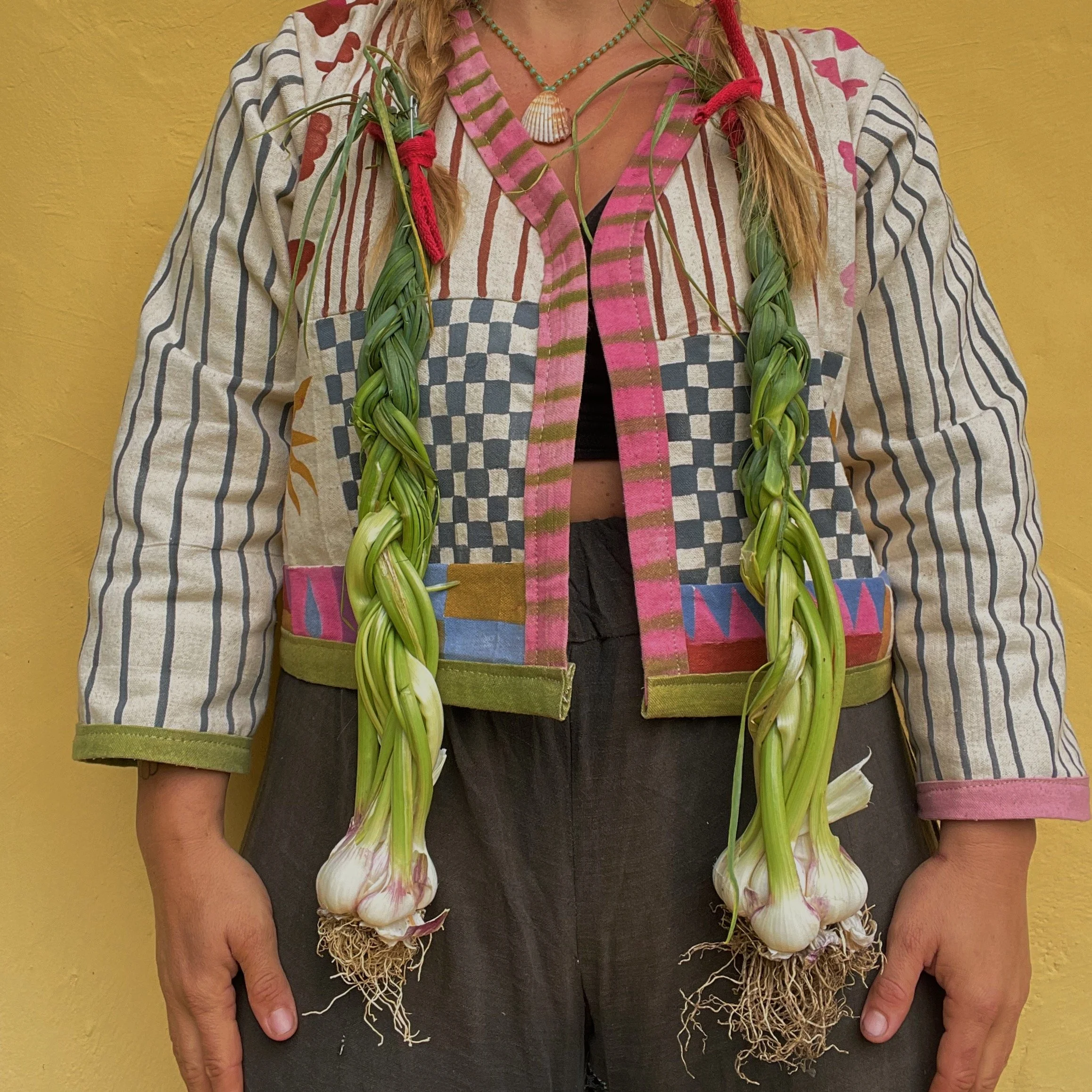 A person wearing a patchwork jacket adorned with garlic bulbs and green plant fibers, a seashell necklace, and a braided braid against a yellow wall.