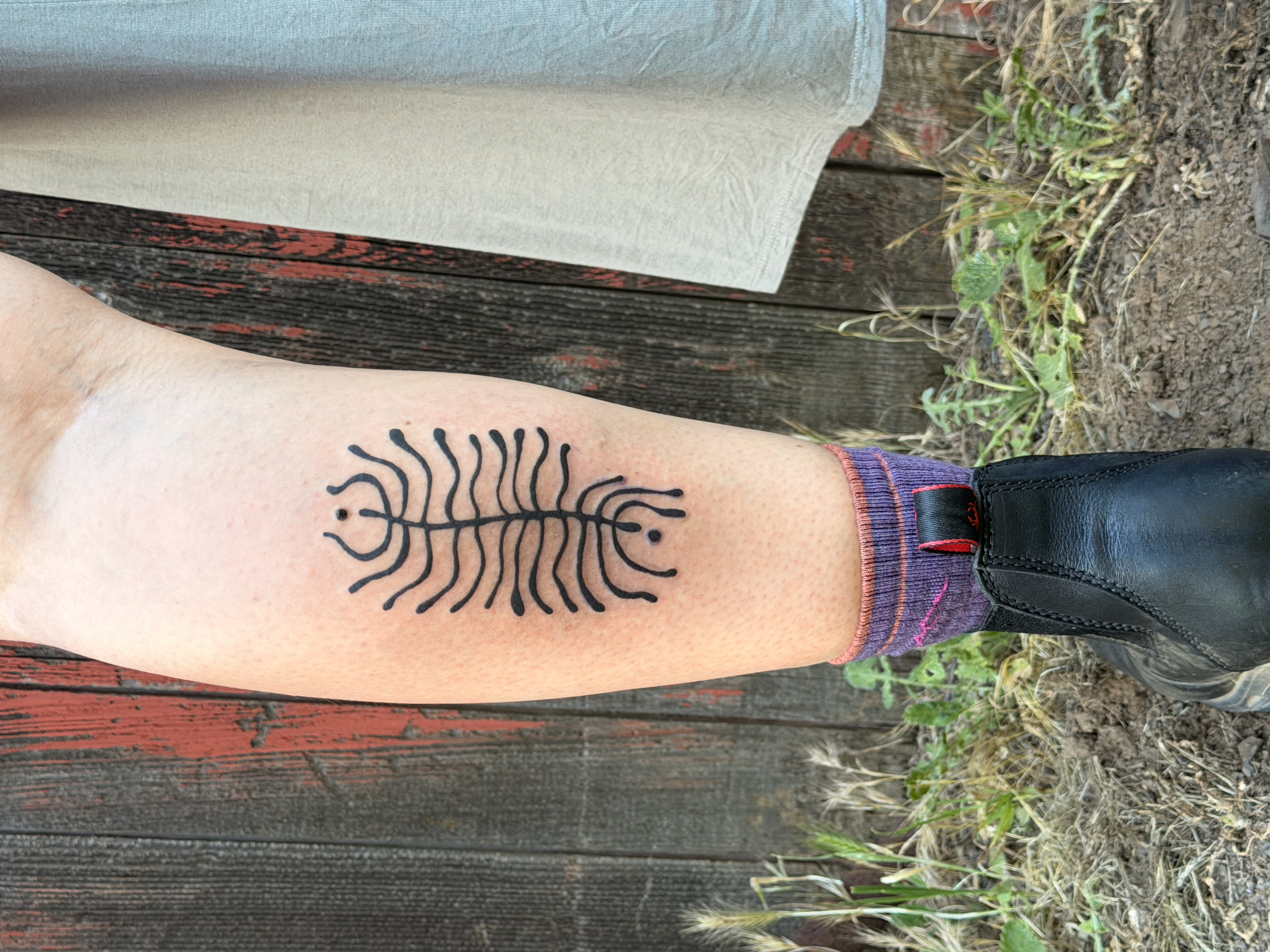 A person's arm with a black tribal-style tattoo of a scorpion lying on a weathered wooden surface next to some small plants.