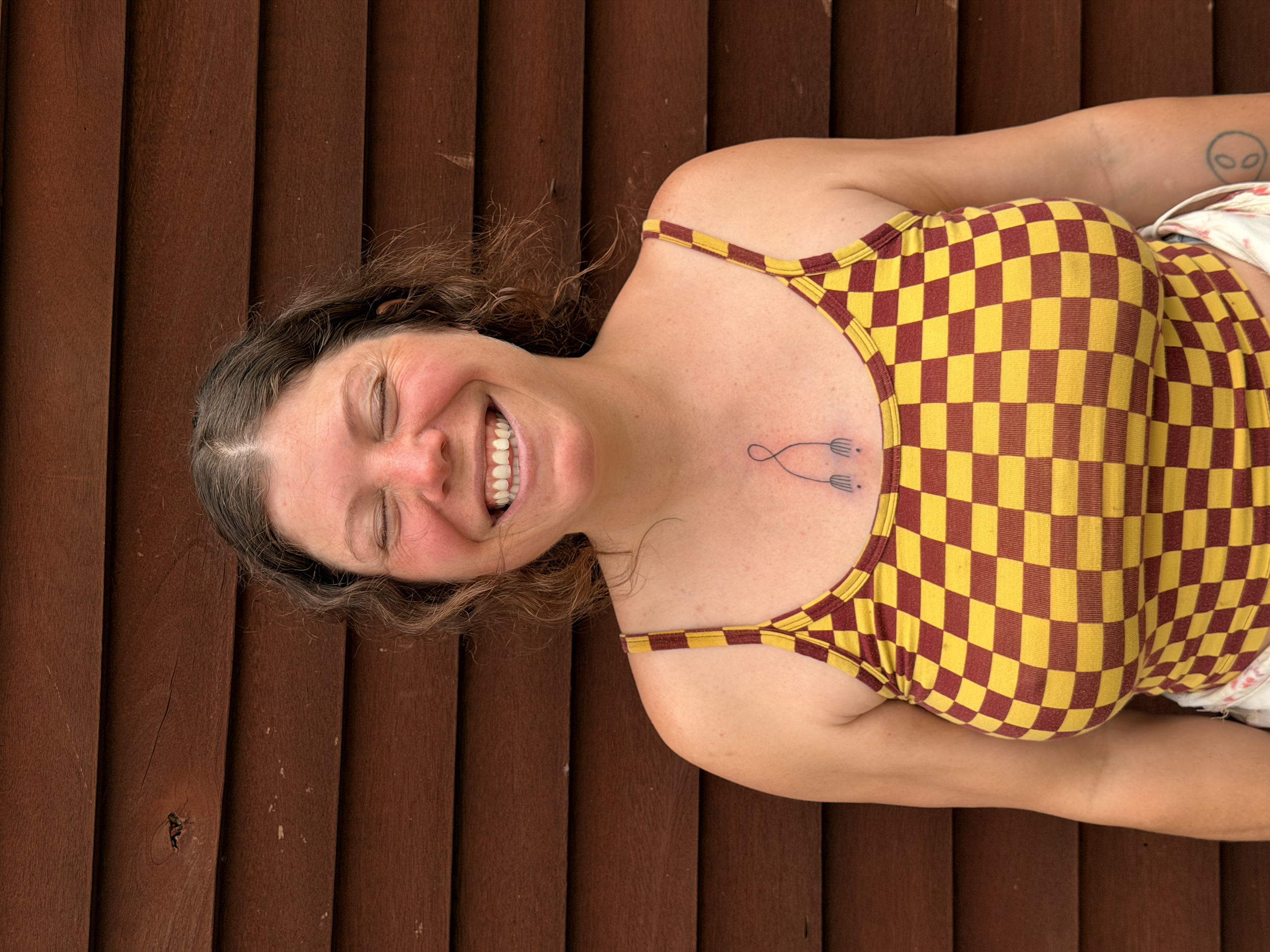 A woman with curly brown hair lying on a wooden surface smiling with eyes closed, wearing a checkered yellow and red dress, a tattoo on her chest, and a tattoo of a face on her arm.