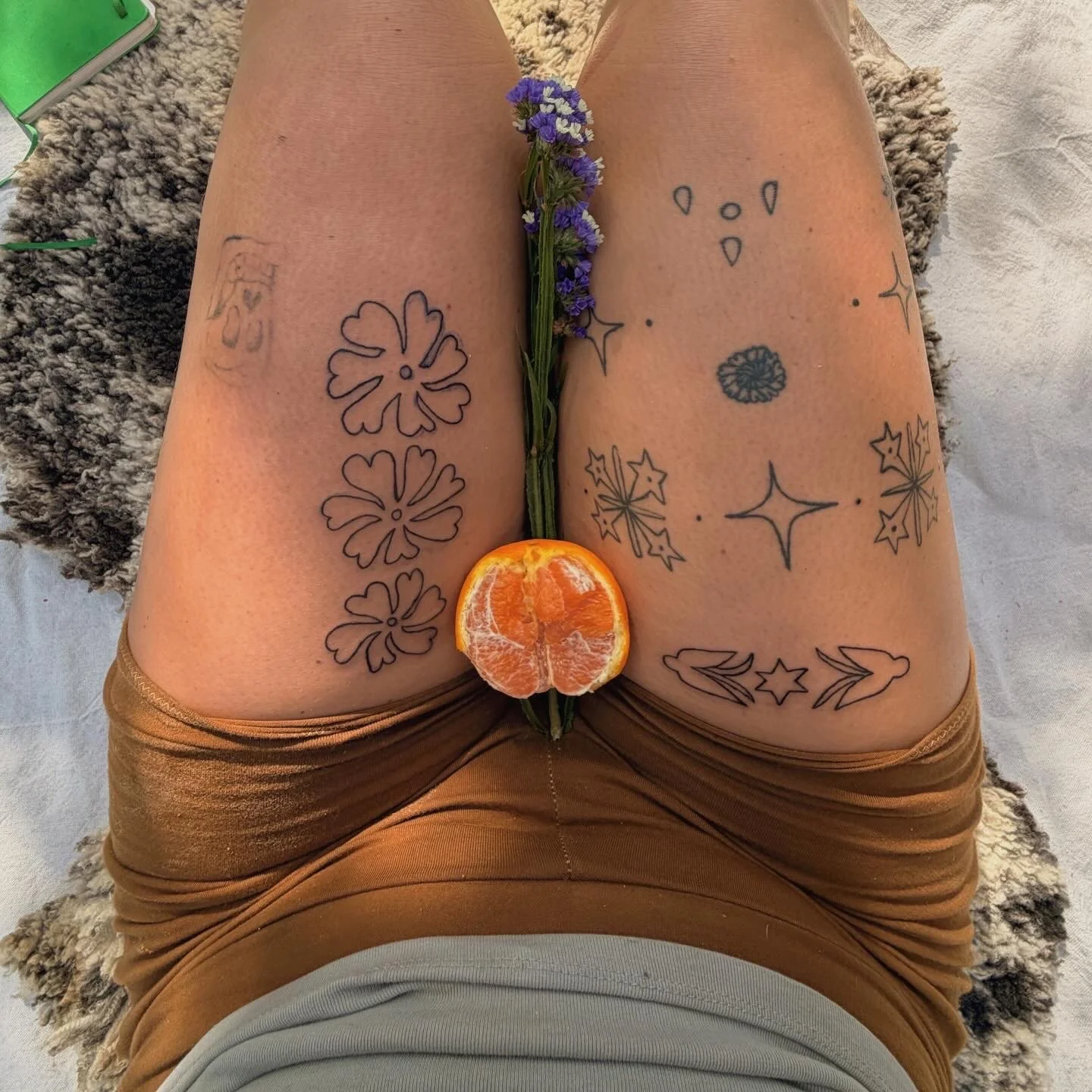 Person lying on their back with tattooed thighs visible, holding a striped orange and a sprig of purple flowers positioned vertically, with the flowers spanning between the thighs. The person is wearing brown shorts and a gray shirt.