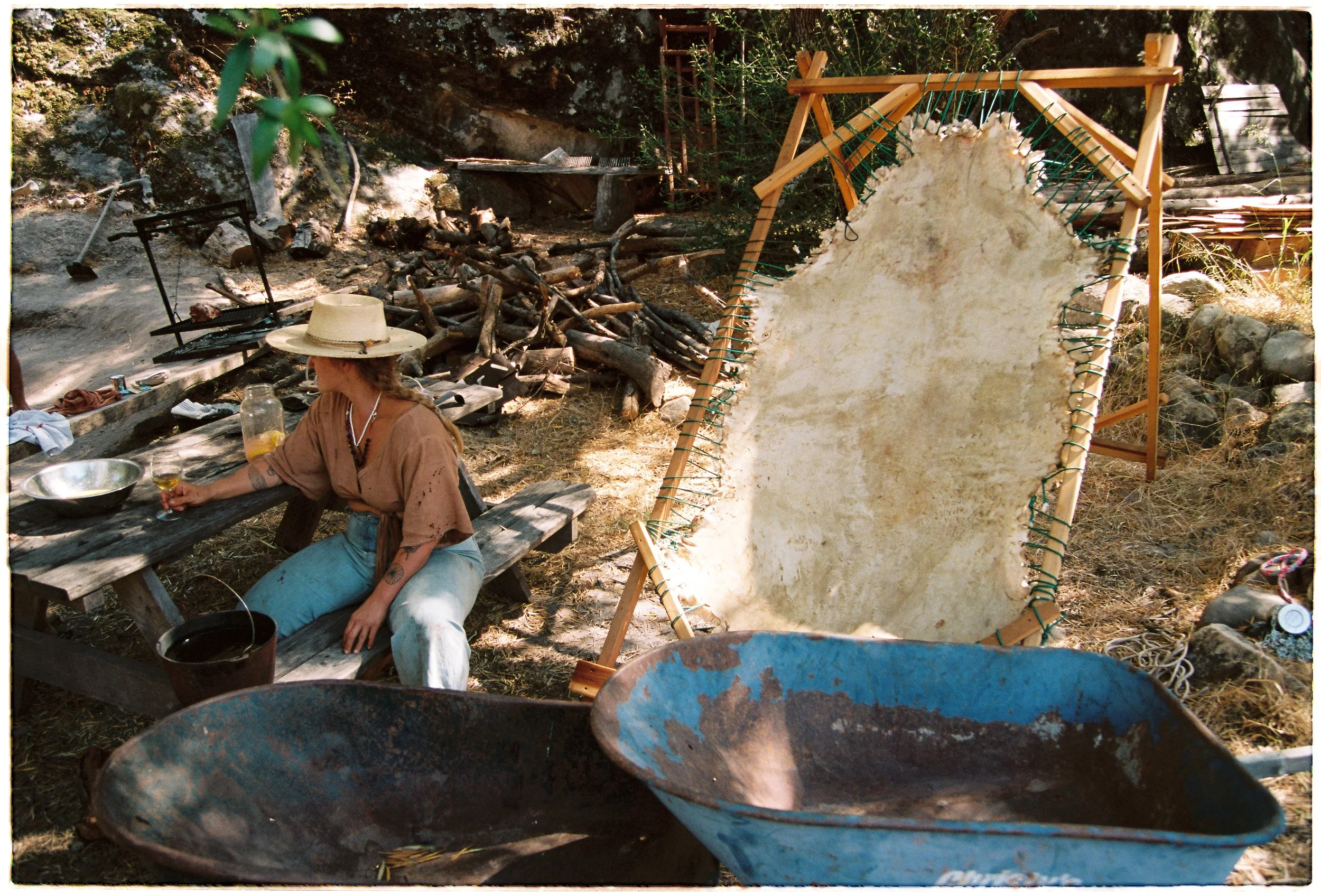 A woman sitting outdoors at a rustic table with a hat on her head, surrounded by wood and equipment used for wool or fiber processing. There is a large homemade loom to the right and a fire pit with logs in the background.