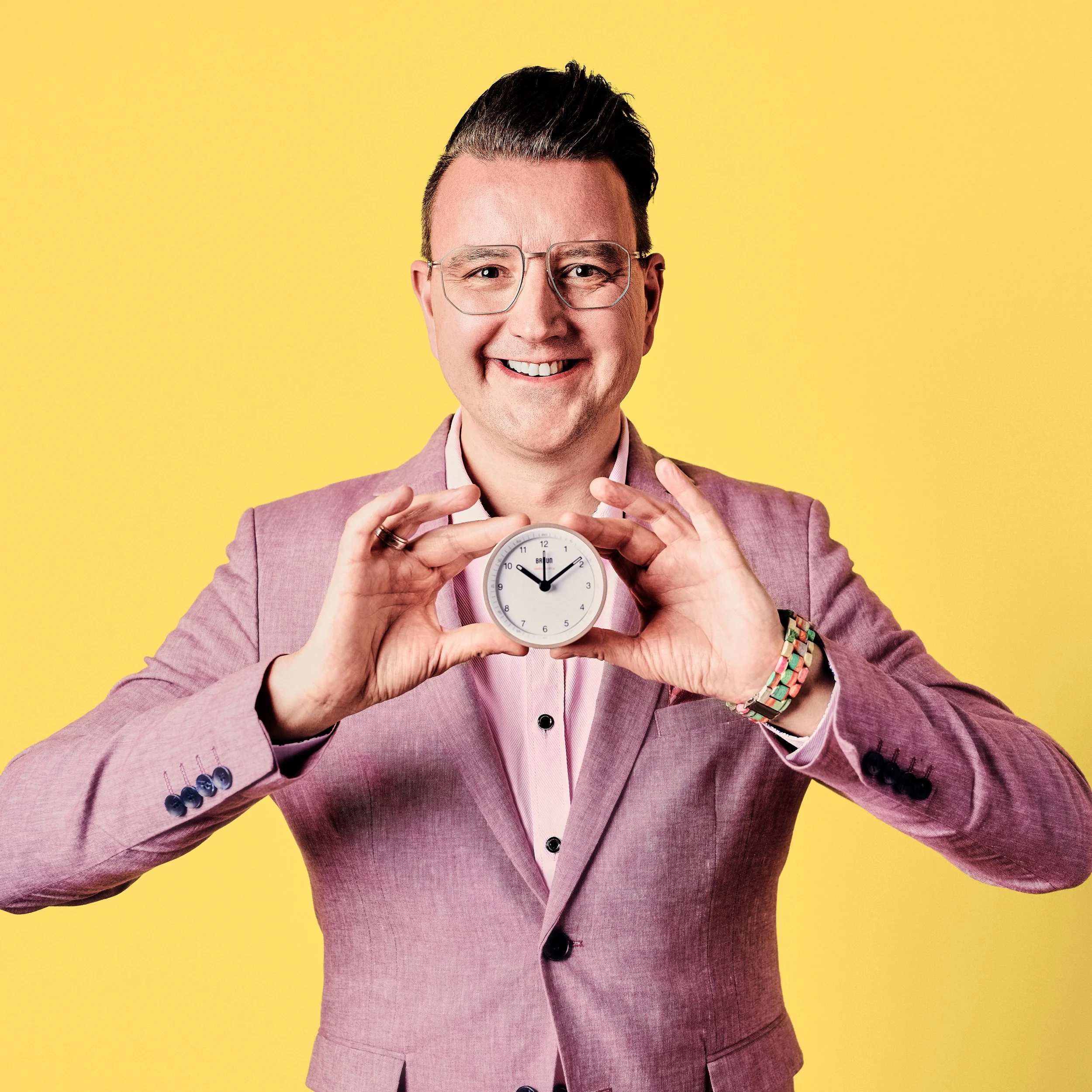 Daniel Winkler lächelt mit Brille und hält eine Wanduhr vor gelbem Hintergrund.