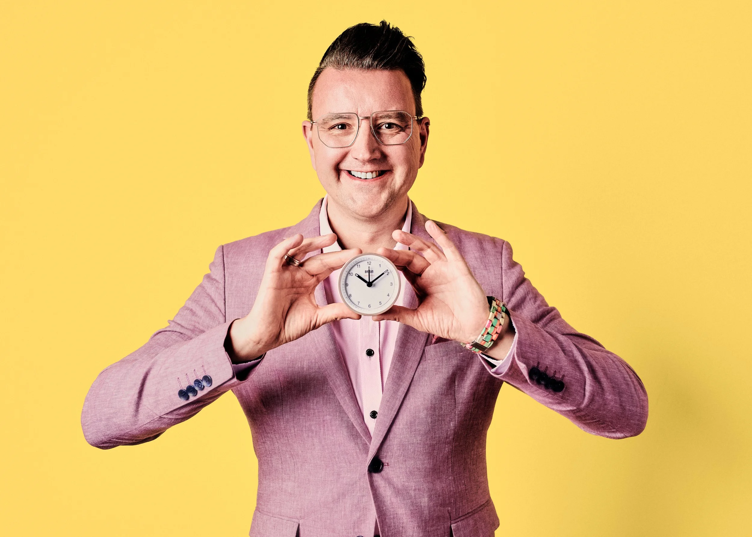 Daniel Winkler in einem rosa Anzug hält eine weiße Uhr vor sich und lächelt vor einem gelben Hintergrund.