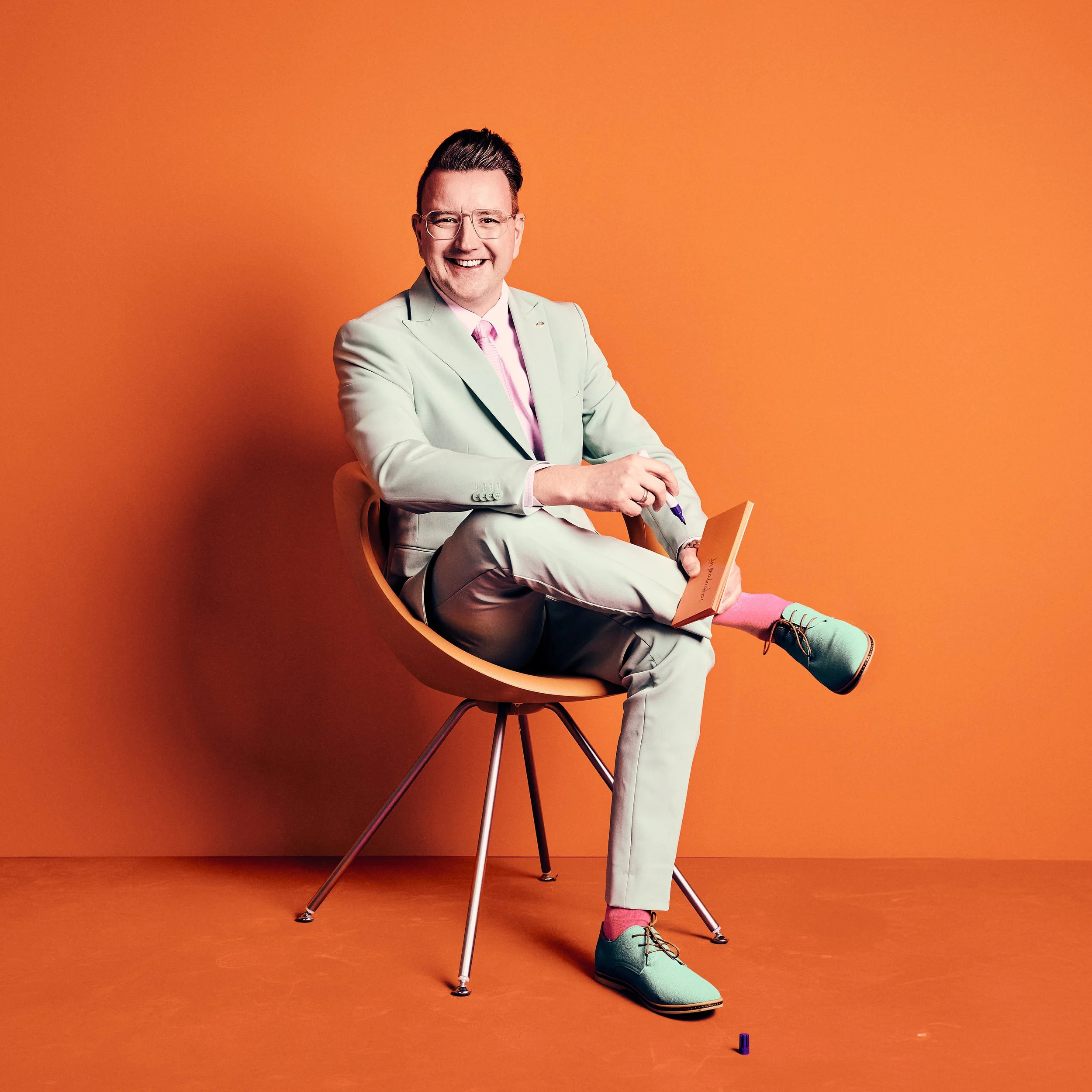 Daniel Winkler lächelt in hellgrauem Anzug, rosa Krawatte, bunten Socken und Brille sitzt auf einem orangefarbenen Stuhl vor einem orangefarbenen Hintergrund, hält einen Marker und liest ein orangefarbenes Buch.