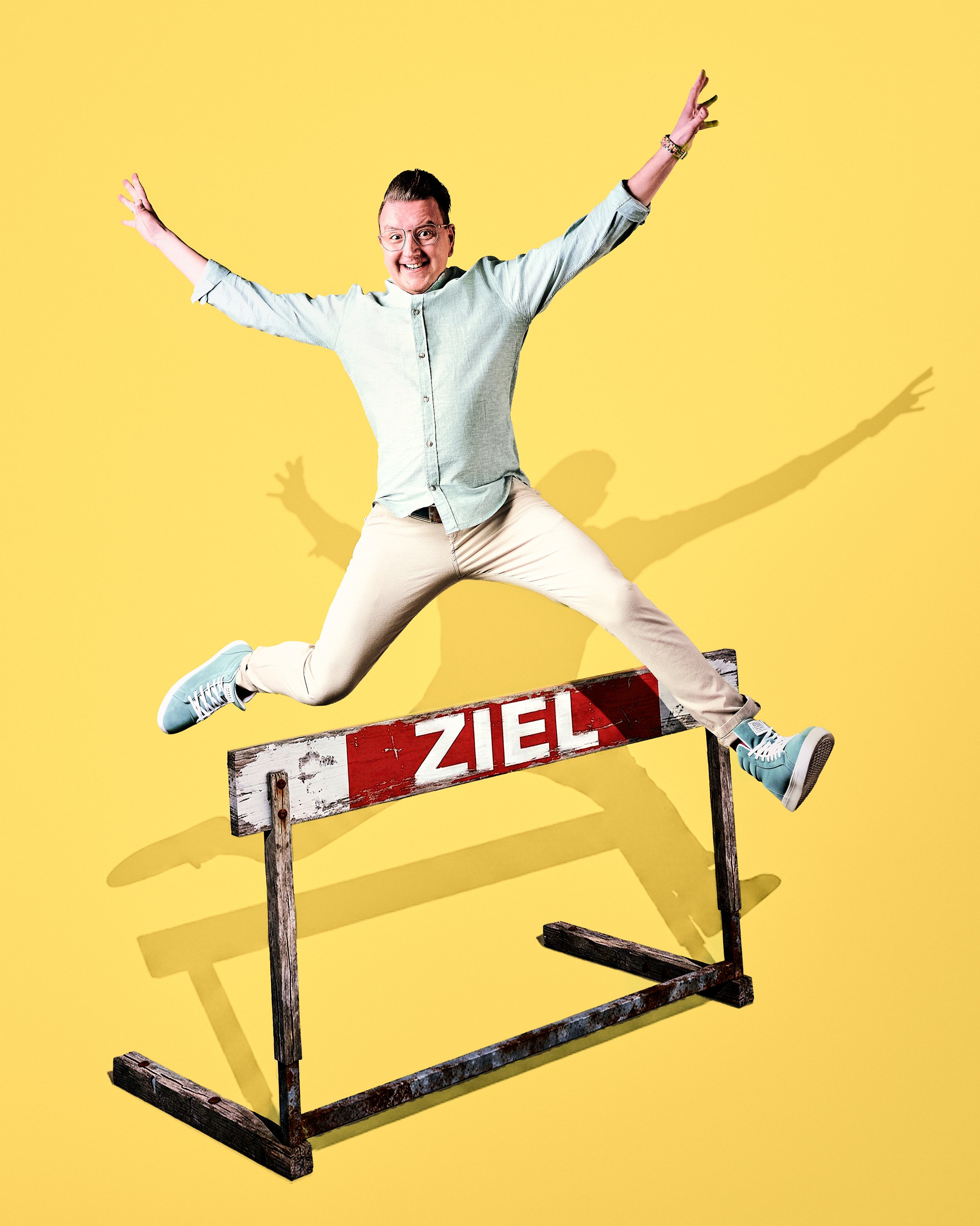Daniel Winkler mit blauer Jacke, beige Hosen und Turnschuhen springt über ein Zielschild mit der Aufschrift 'ZIEL' vor einem gelben Hintergrund. Seine Schatten sind deutlich sichtbar.