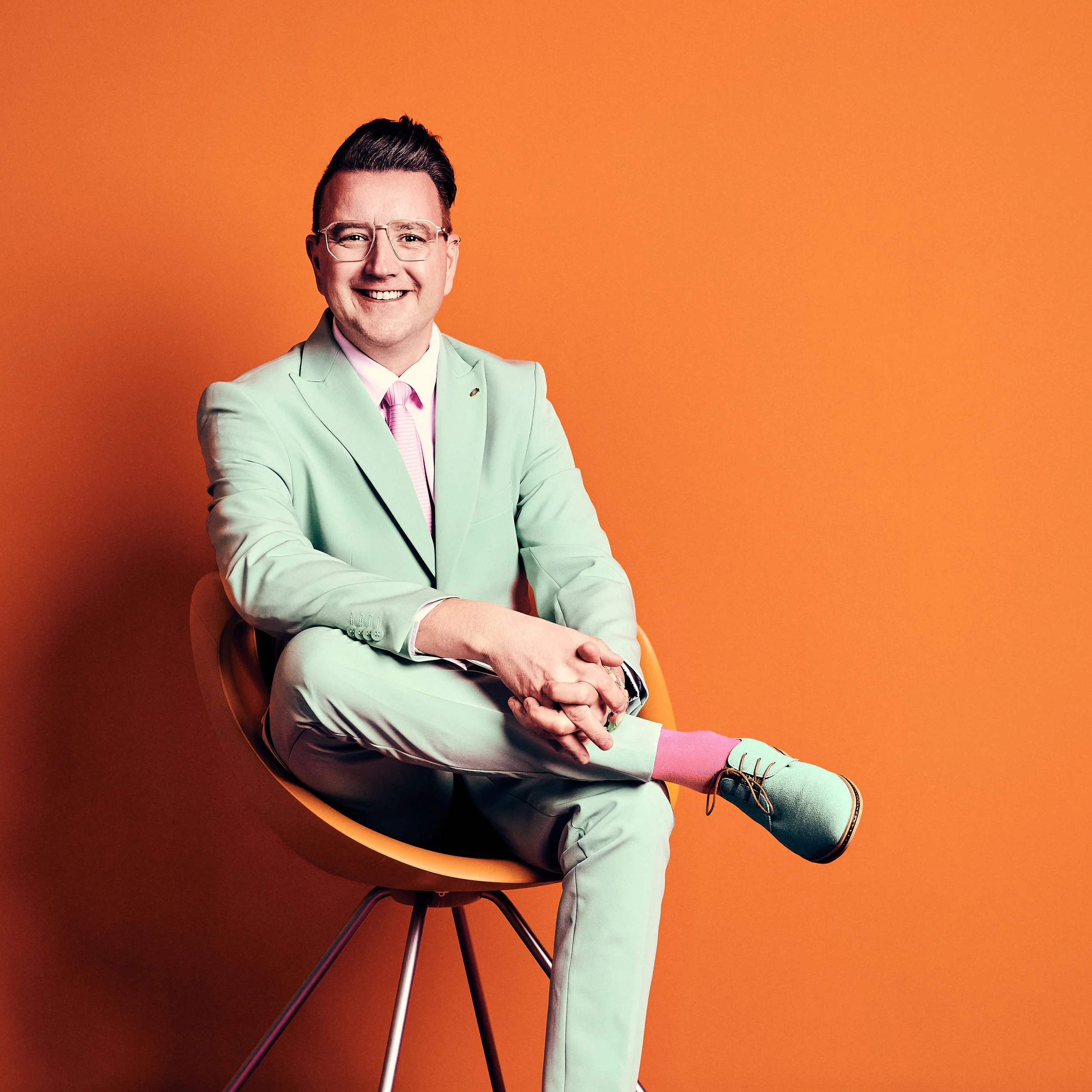 Daniel Winkler mit Brille, grünem Anzug, pinkem Krawattenknoten, sitzend in einem orangefarbenen Sitz vor einem orangefarbenen Hintergrund.