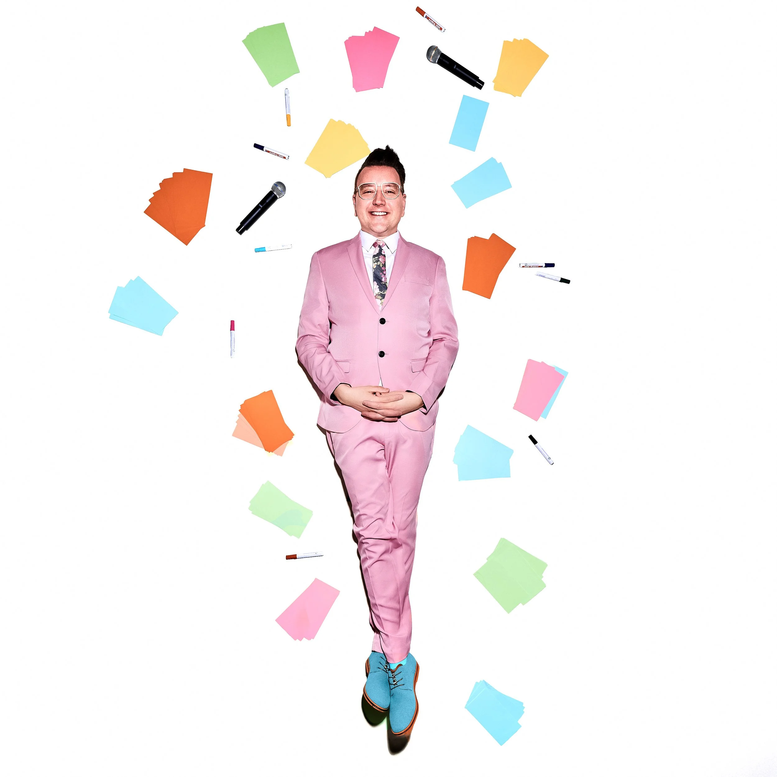 Daniel Winkler in einem pinken Anzug mit einer Krawatte steht vor weißem Hintergrund, umgeben von bunten Papierbällen, Filzstiften, Markern, einem Mikrofon und Haftnoten, lächelnd und fröhlich.