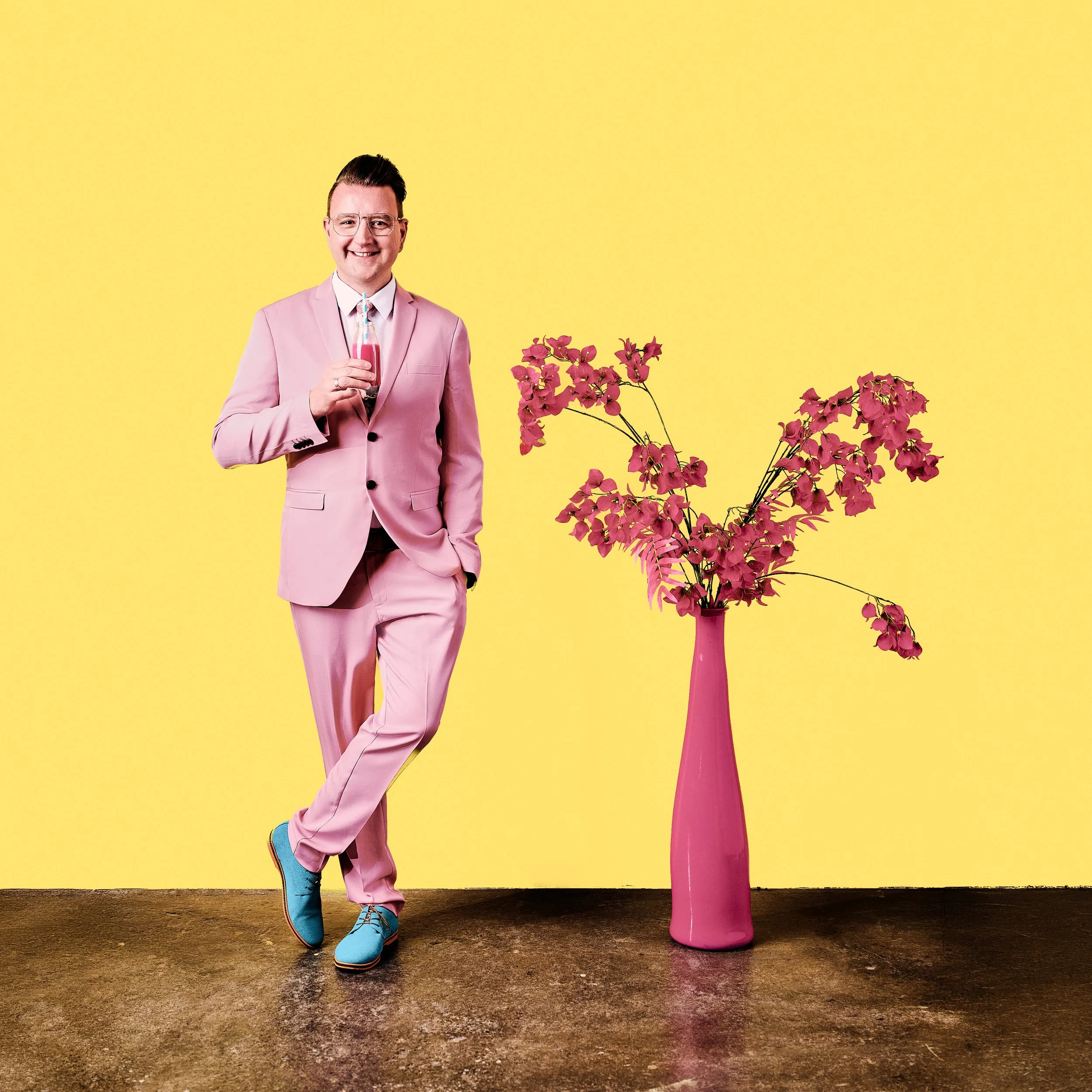 Daniel Winkler in Pinkem Anzug, Brille und blauen Schuhen lächelt, hält ein Glas mit rotem Getränk, steht neben einer großen pinken Vase mit roten Blumen, vor einem gelben Hintergrund.