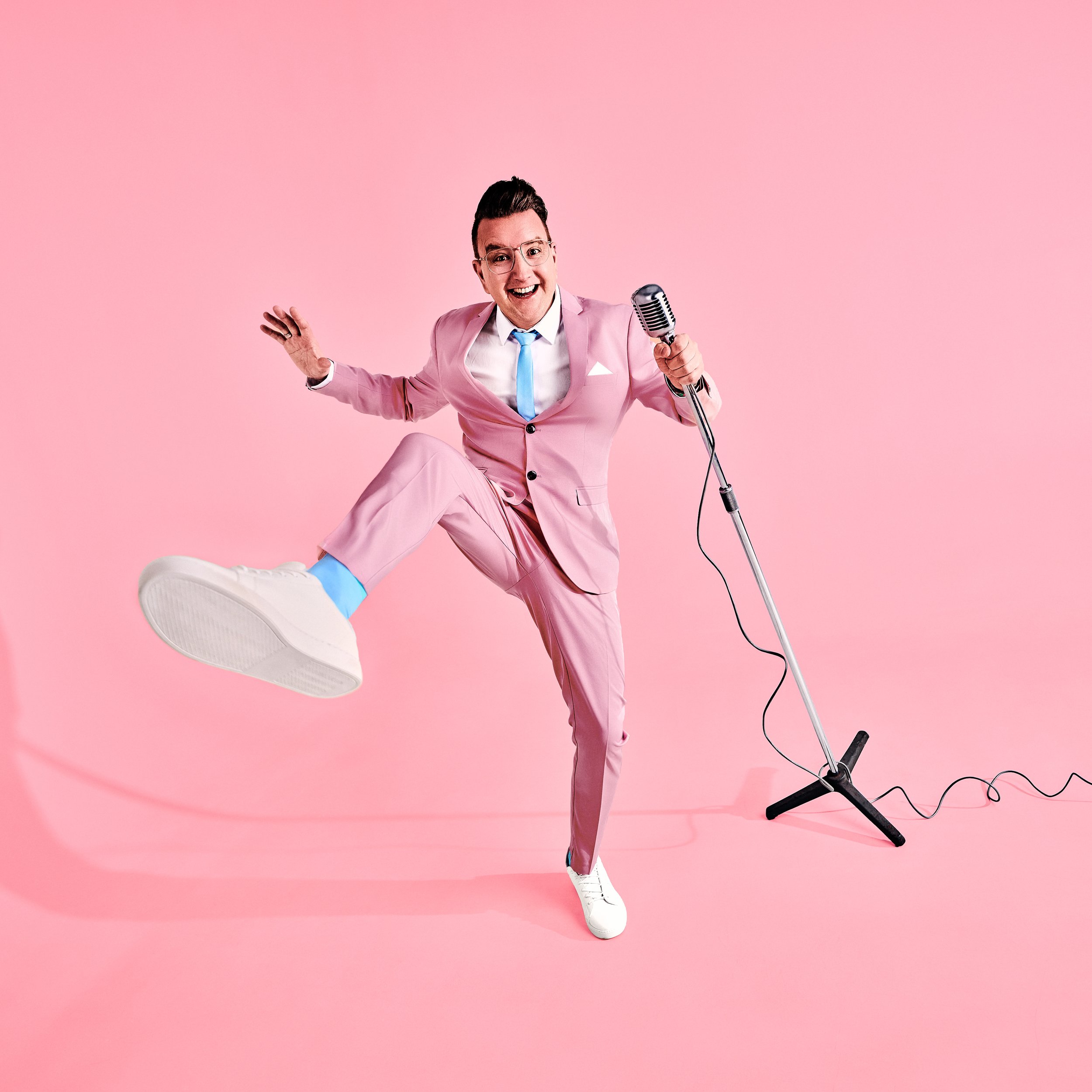 Daniel Winkle in rosa Anzug mit blauer Krawatte hält ein Mikrofon vor pinkem Hintergrund, hebt eine Hand und hat einen Bein hochgestreckt.