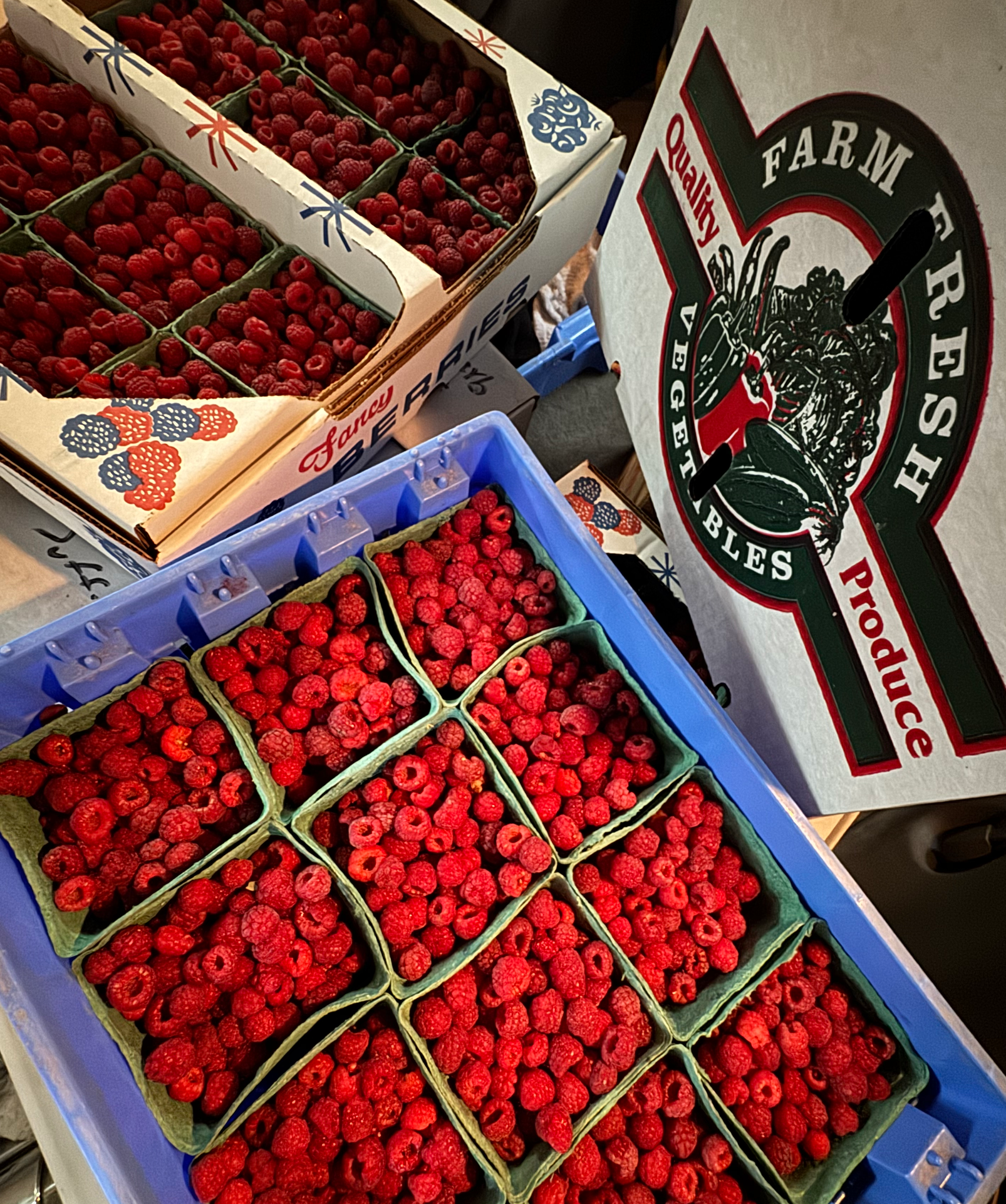 Boxes of fresh red raspberries on display at a market, with a farm produce sign visible nearby.