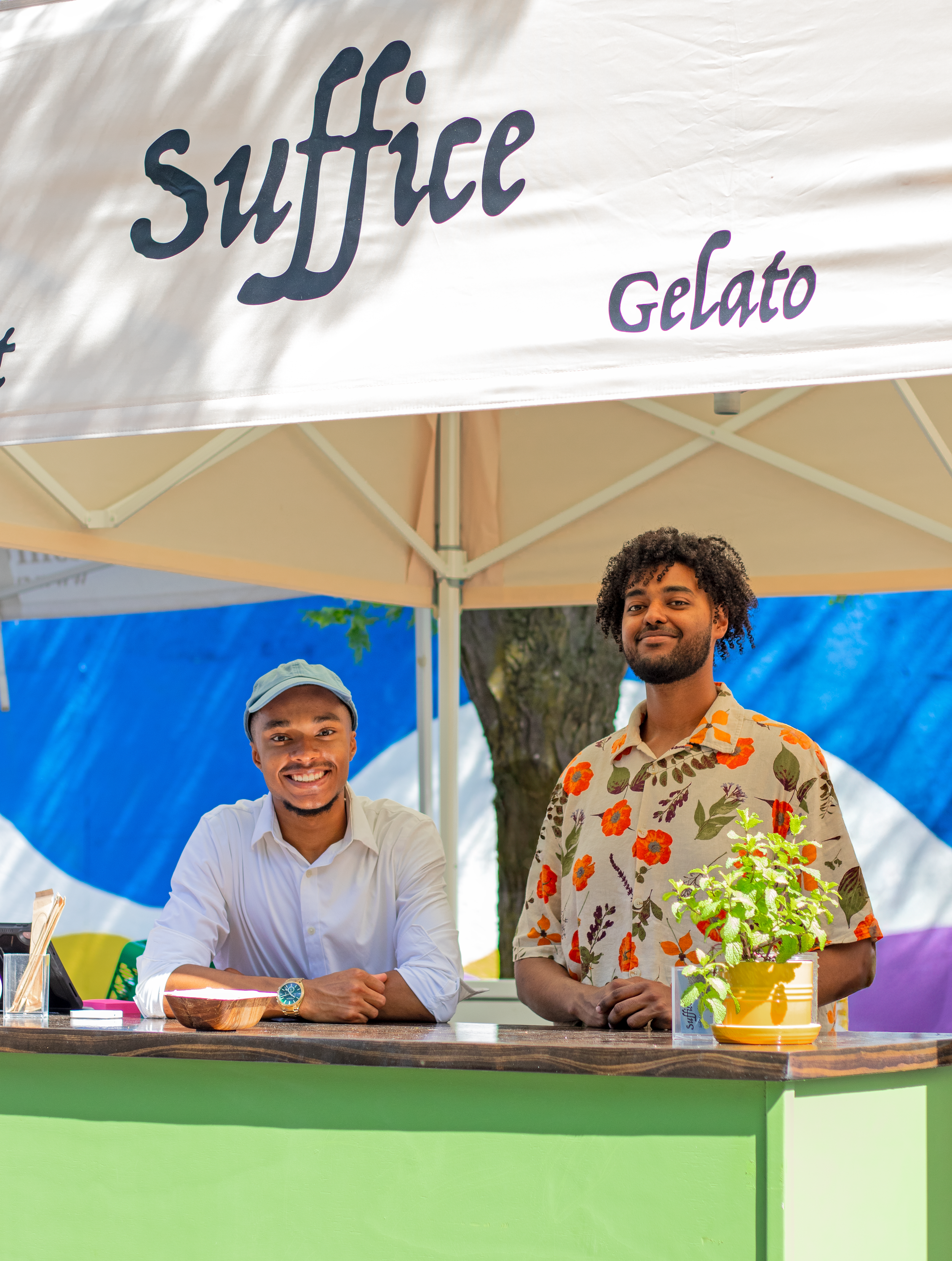 Two men standing behind a green booth with a potted herb on the table. One man is seated and wearing a white shirt and watch, smiling, while the other man is standing, wearing a floral shirt, smiling. A sign reads "Suffice Gelato."