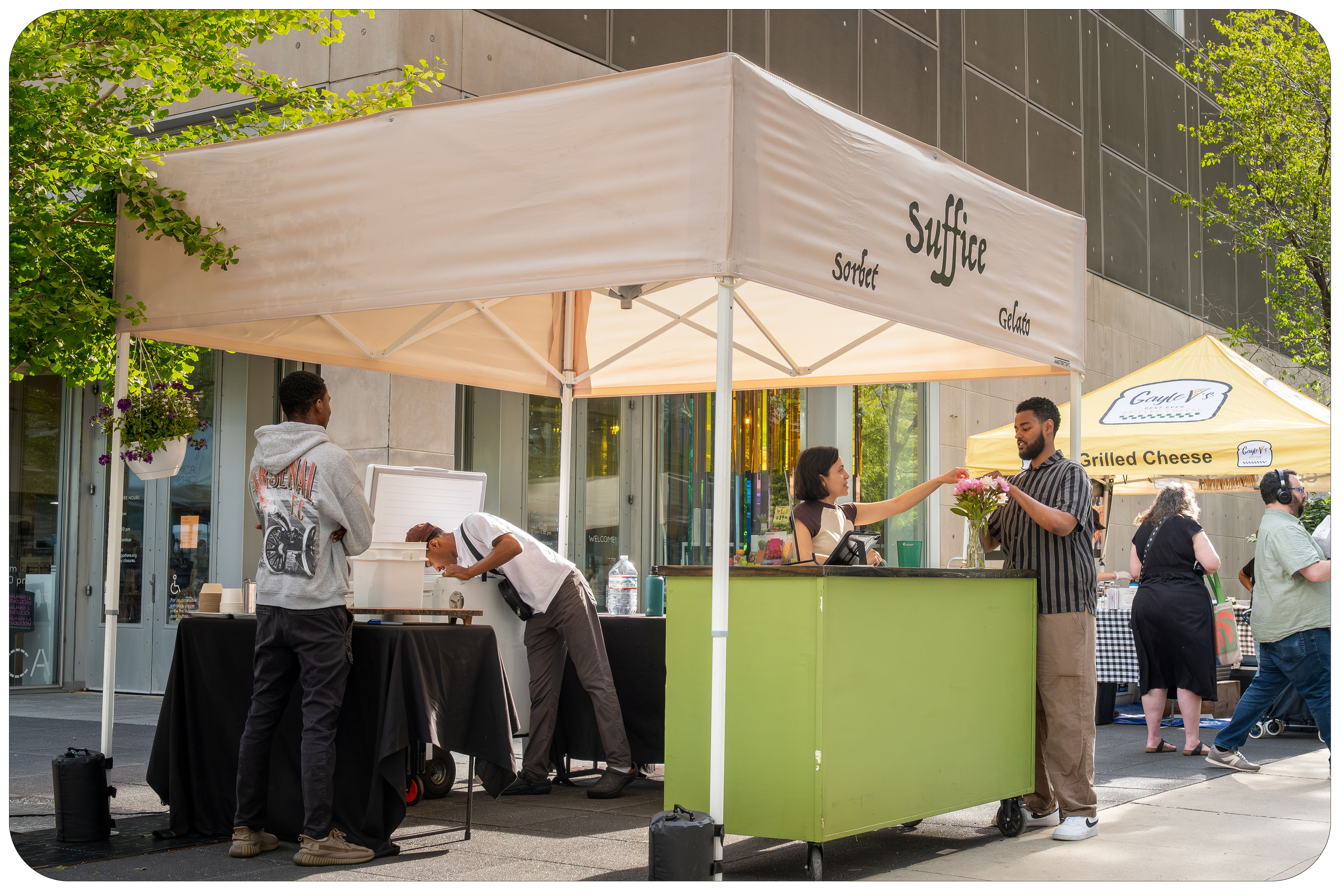 Outdoor marketplace stand with a beige canopy labeled 'suffice' selling sorbet, gelato, and grilled cheese. People are gathered around, with some ordering and others preparing items, in front of glass-fronted buildings and surrounded by trees.