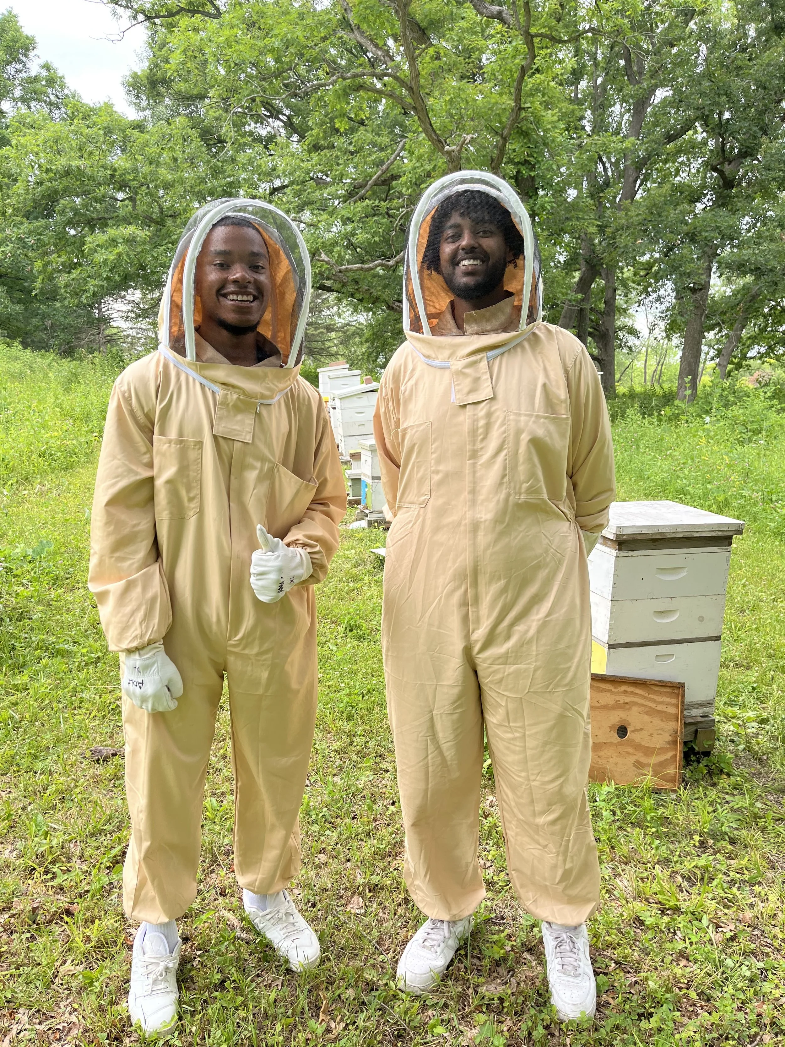 Two men in beekeeping suits standing in a grassy outdoor area with beehives behind them and trees in the background.