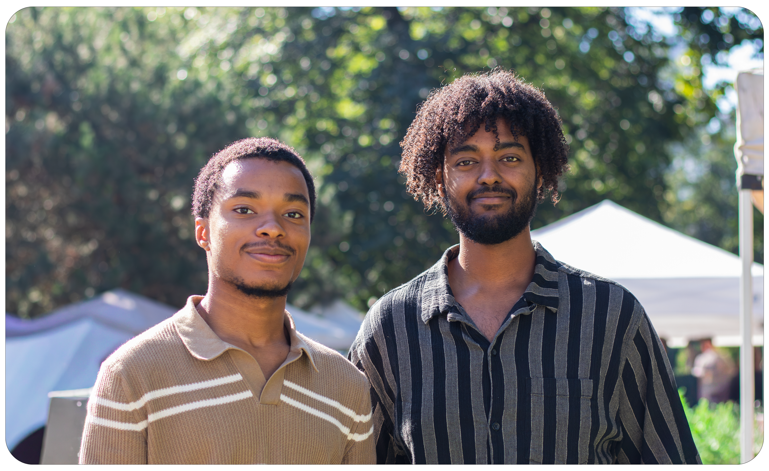Two young men standing outdoors with trees in the background, smiling at the camera. One is wearing a beige and white striped polo shirt, and the other is wearing a black and gray striped shirt.