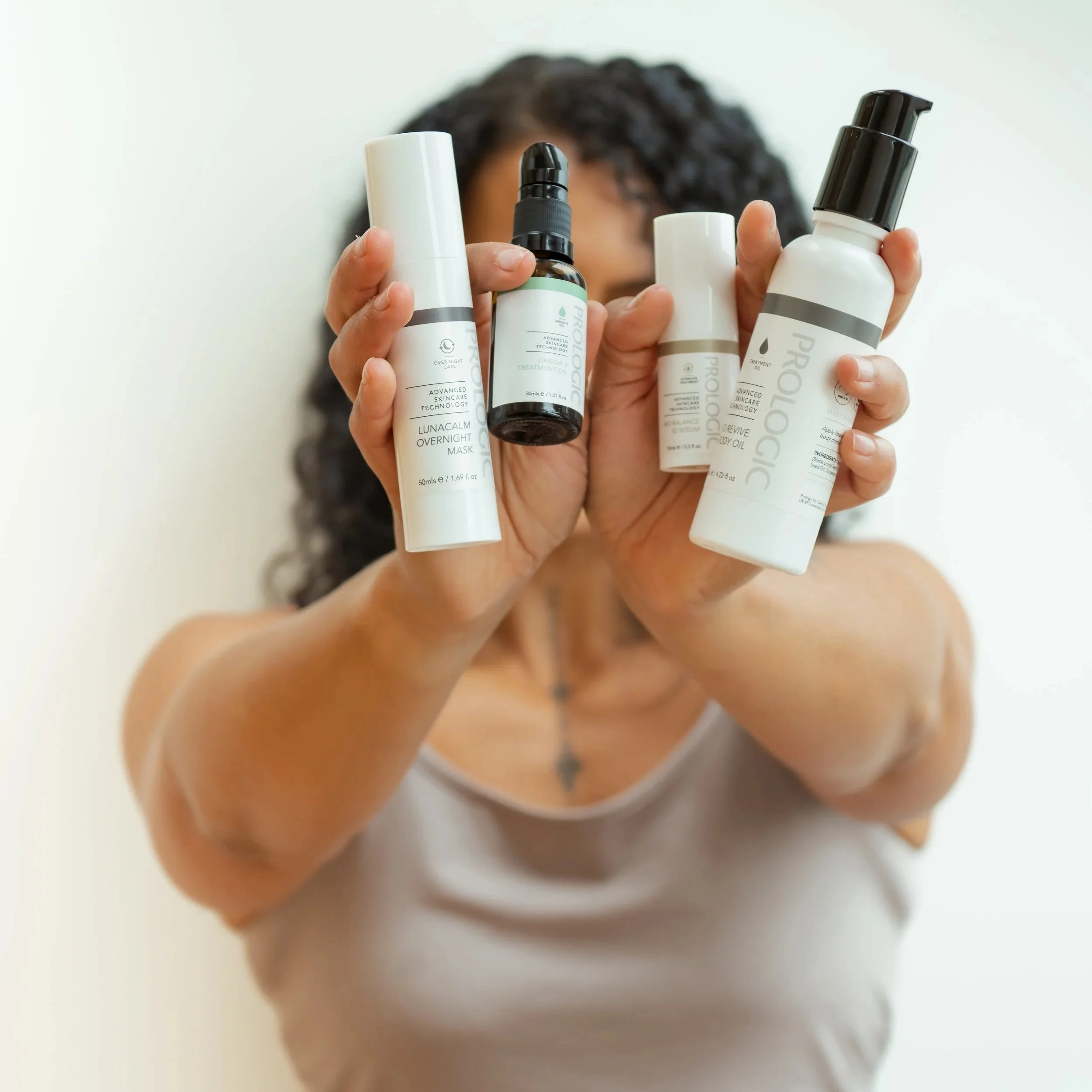 A person with curly dark hair holding four skincare bottles and a dropper in front of their face, obscuring their facial features, against a plain light background.