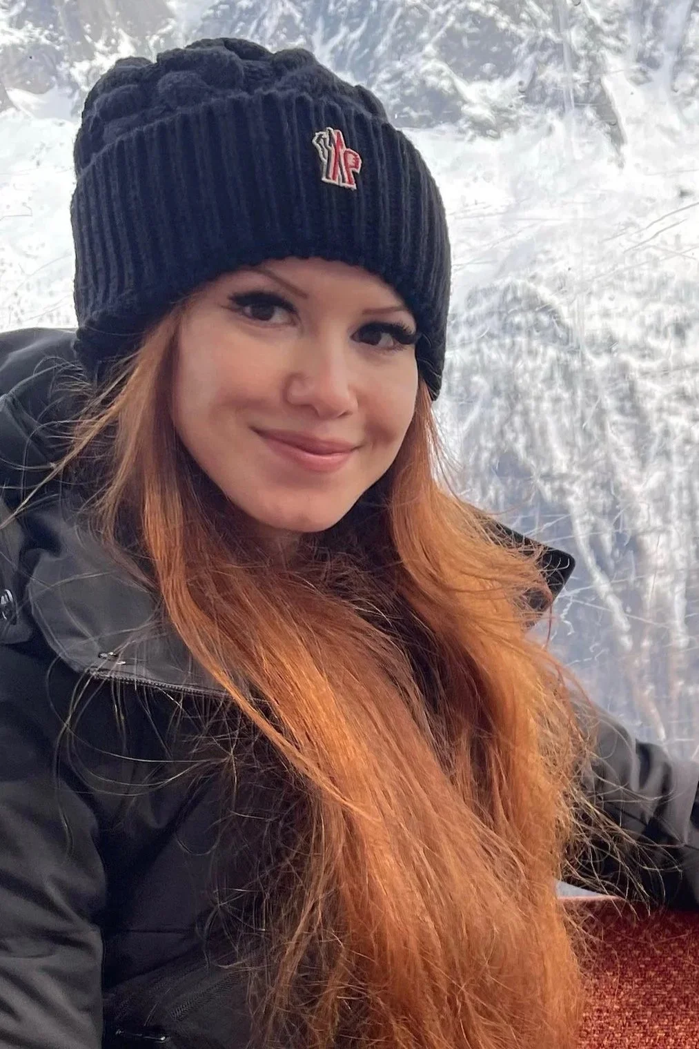A woman with long red hair smiling, wearing a black knit beanie and a black jacket, against a snowy background.