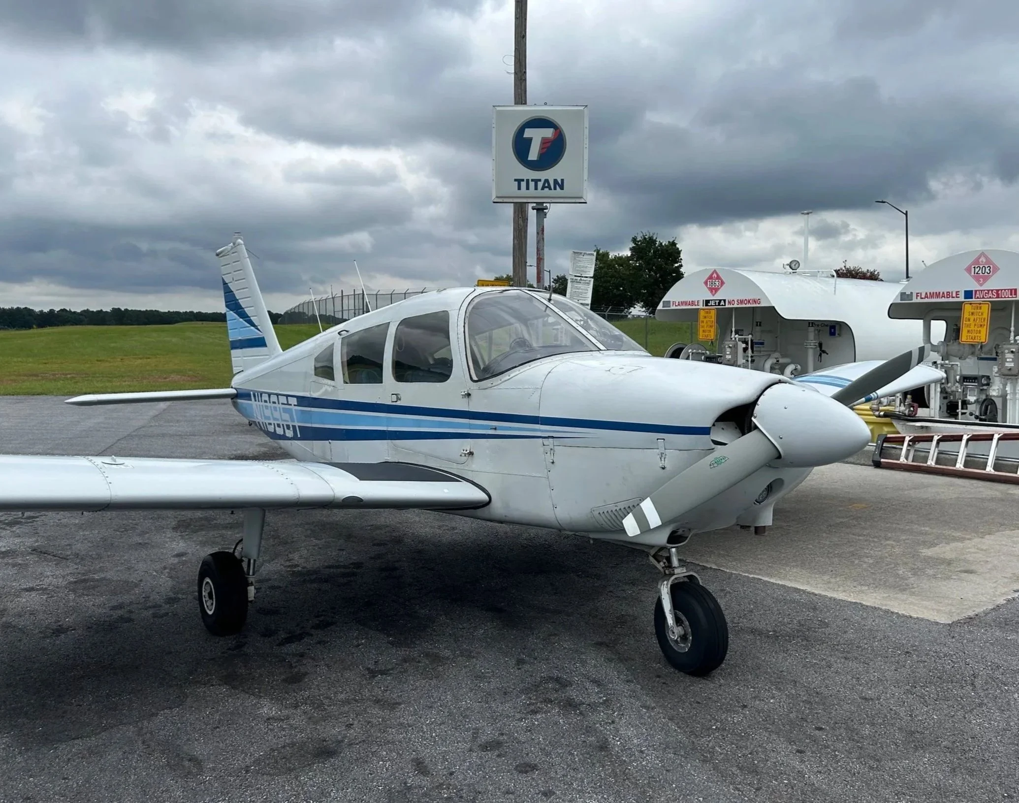 A small white and blue aircraft parked on a tarmac, with fuel tanks and a fueling station in the background under a cloudy sky.