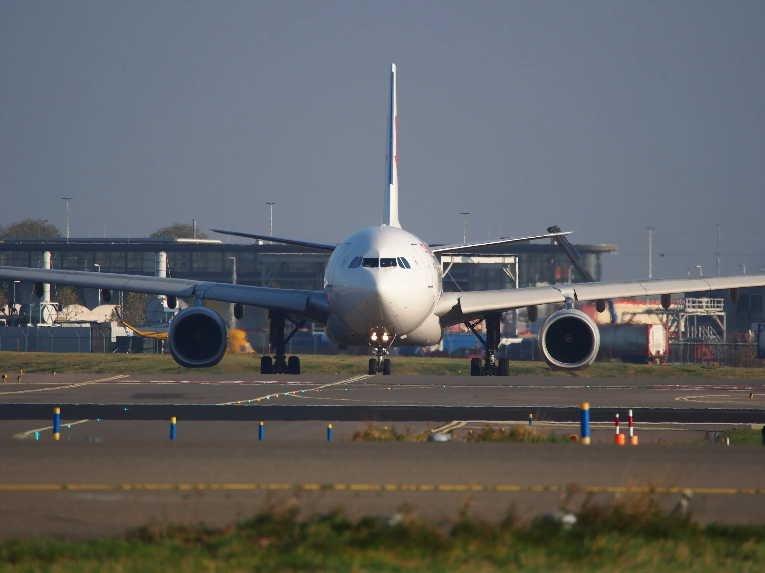 Front view of a commercial airplane on the tarmac at an airport, with the airport terminal and hangars in the background, under a clear sky.