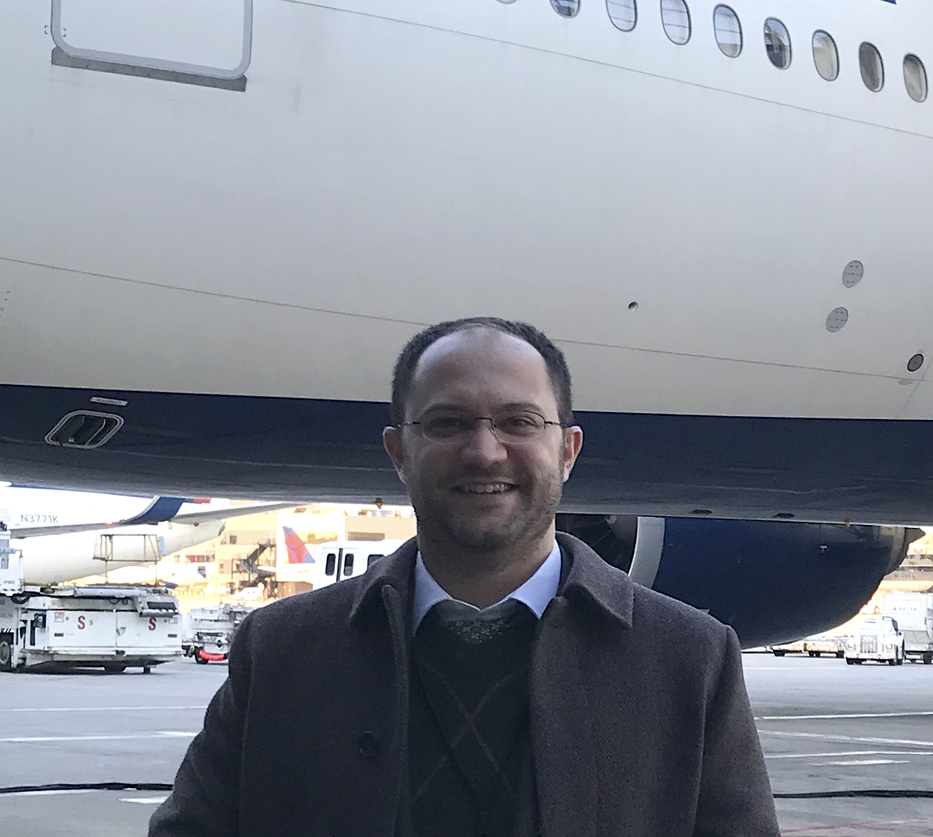 Attorney Mahmoud Khatib, in a wool overcoat, smiles on an airport tarmac with a large airline jet behind him and airport vehicles in the background.
