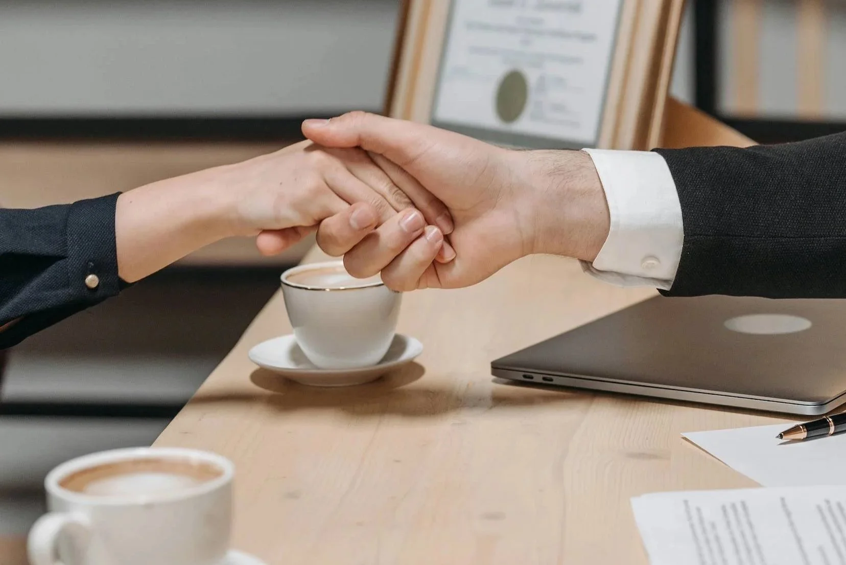 Two people shaking hands over a desk with a laptop, a cup of coffee, and a pen, possibly in a professional or business setting.