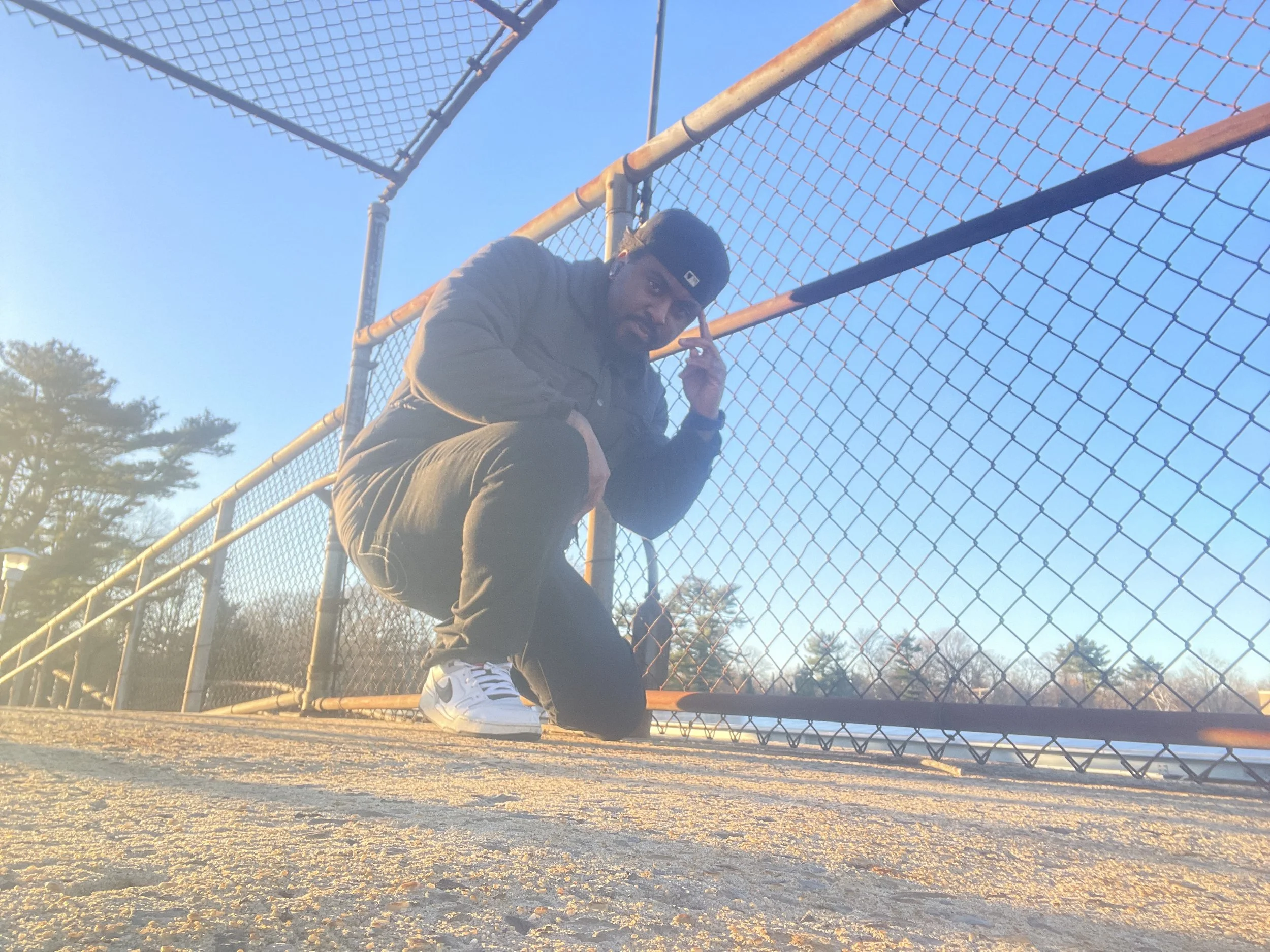 A man dressed in dark clothing, wearing a black beanie, crouching in front of a chain-link fence during the daytime. The sky is clear and blue, and some trees are visible in the background.