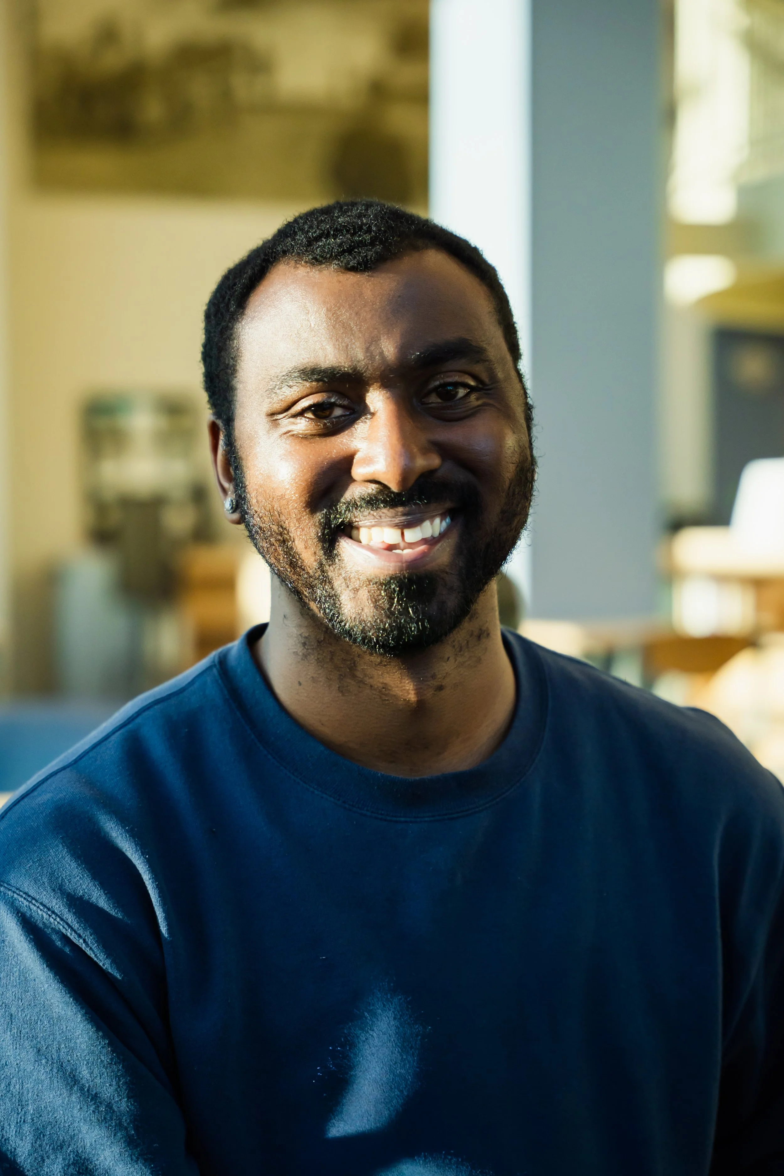 A smiling man with dark curly hair and a beard, wearing a navy blue shirt, is indoors near a window with sunlight.