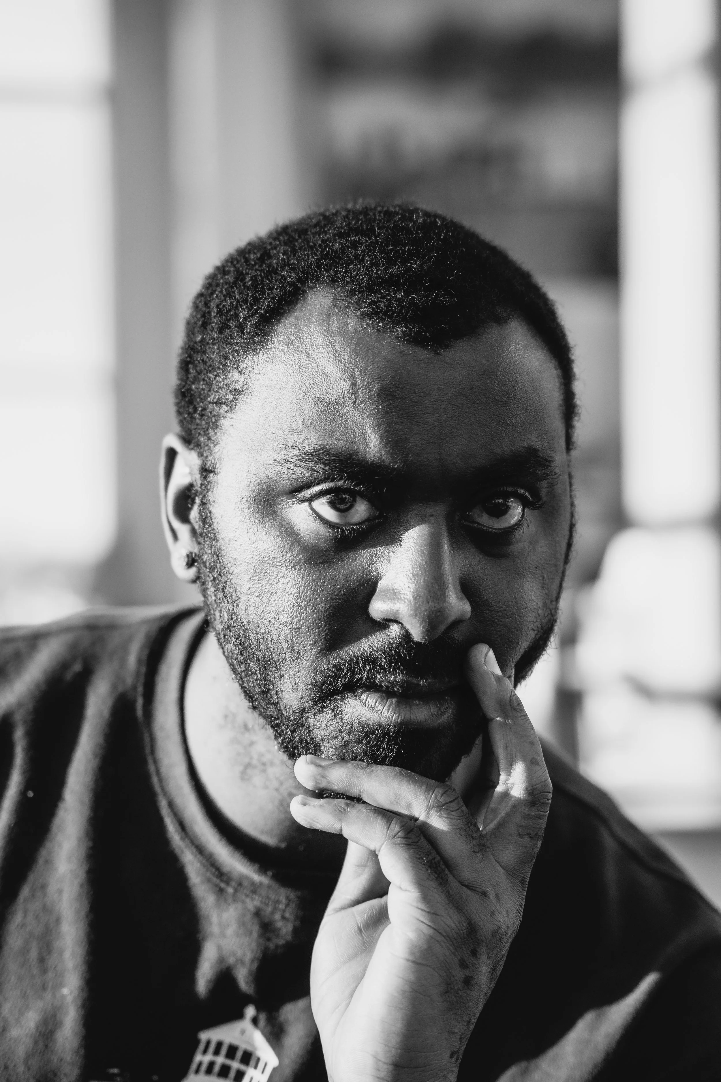 A black and white portrait of a man with short curly hair, a beard, and intense eyes. He is holding his chin with his left hand, with his fingers near his lips, and looking directly at the camera.