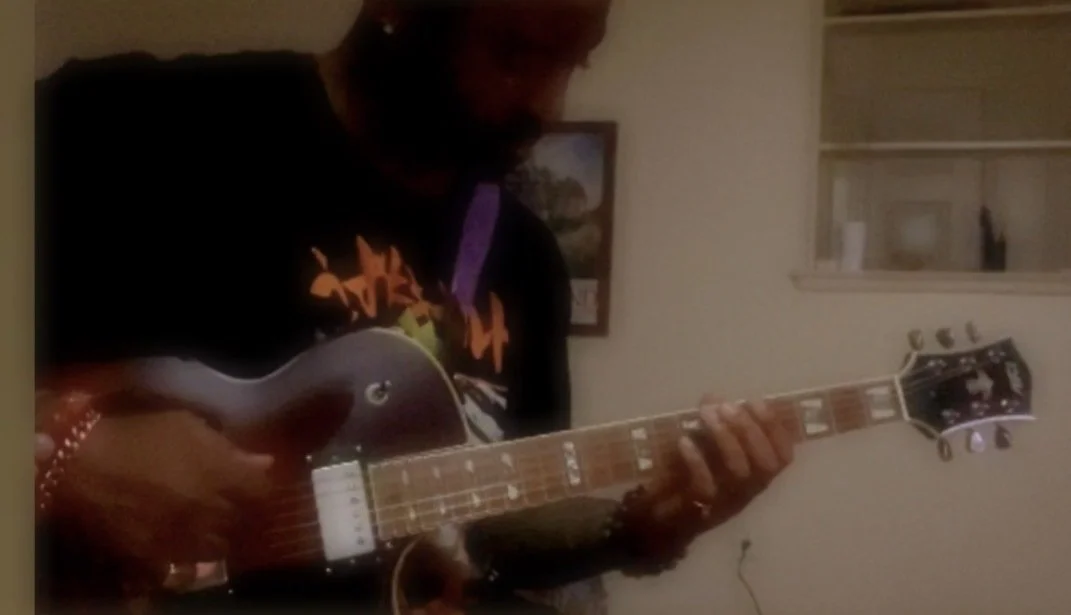 Person playing an electric guitar in a room, with a picture frame on the wall and a window with blinds in the background.