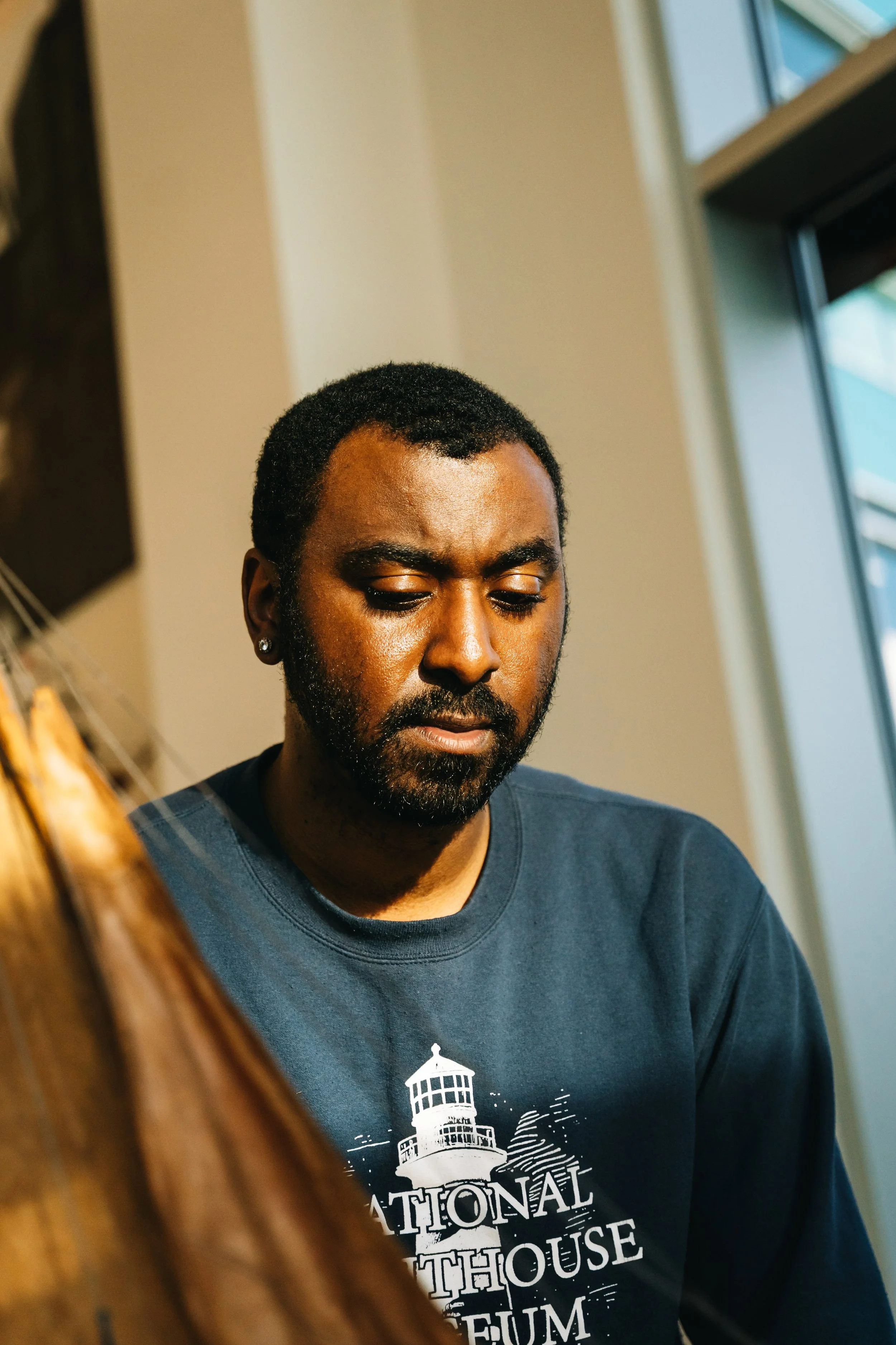 A man with short curly black hair and a beard looking down, wearing a dark blue T-shirt with a lighthouse graphic and text. The background includes a beige wall and a window with glare. Part of a wooden object is visible in the foreground.