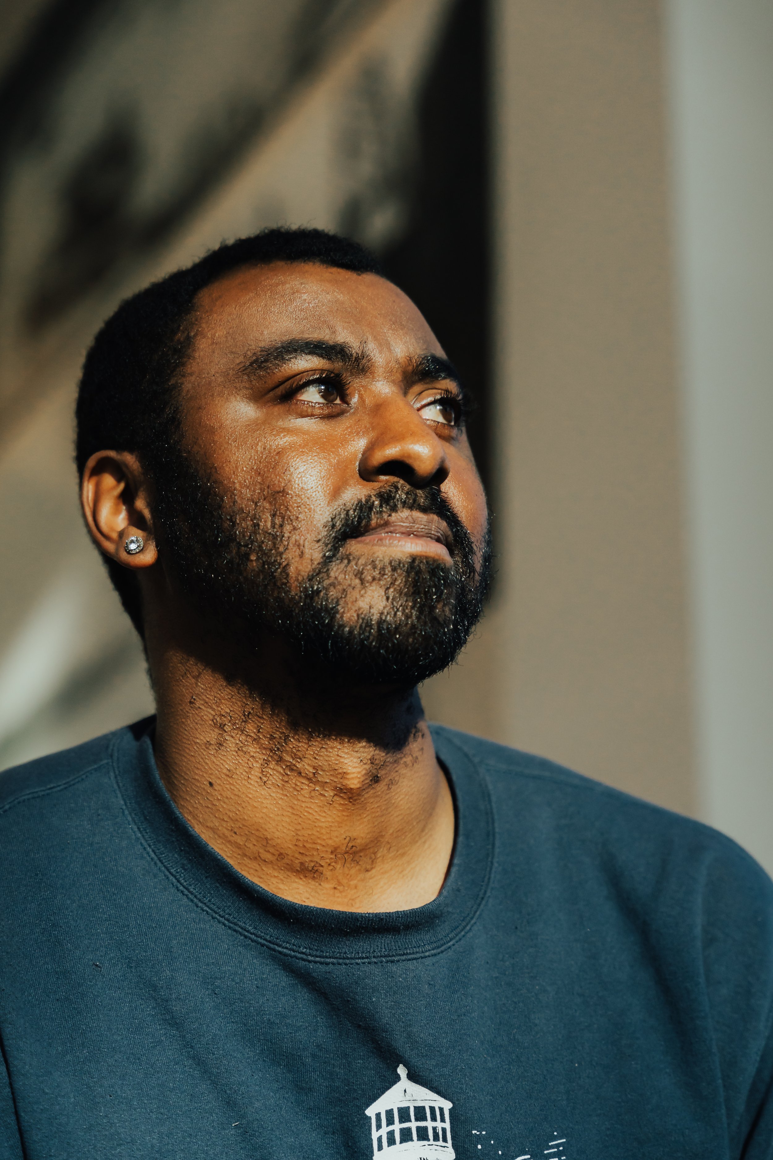 Close-up of a man with dark skin, black hair, a beard, and an earring in his left ear, looking slightly upwards. He is wearing a dark blue T-shirt with a white graphic of a building with columns and a dome.