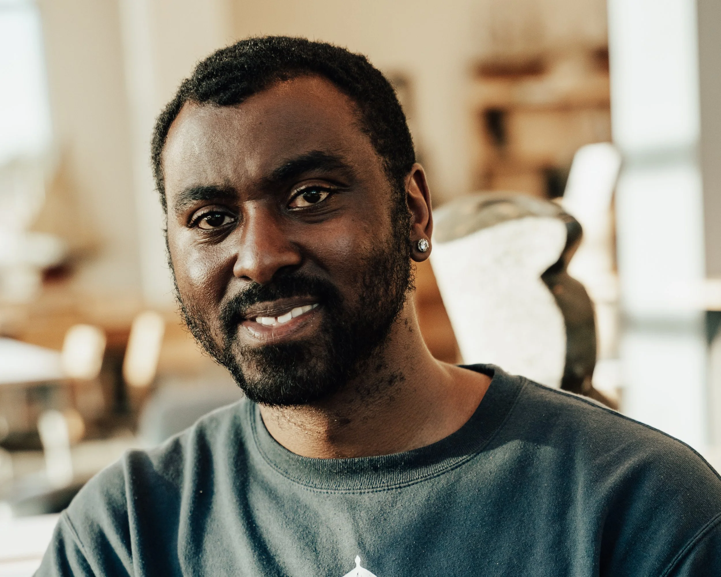 A young African American man smiling at the camera with a beard, wearing a black T-shirt and a small diamond earring in his left ear, indoors with blurred background.
