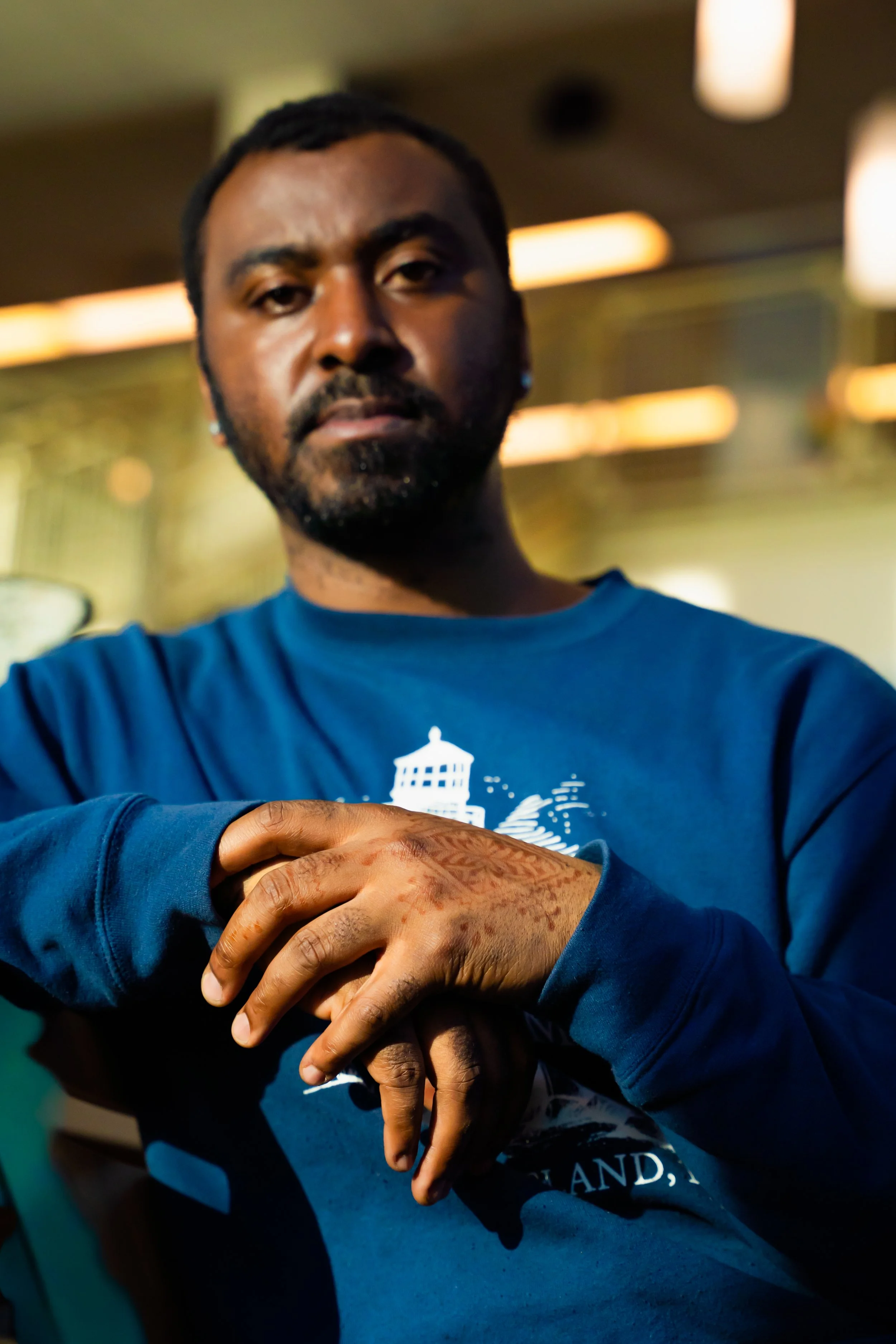 Close-up of a man with dark skin, facial hair, and short dreadlocks wearing a blue long-sleeve shirt with a white graphic, crossing his arms in a dimly lit environment with warm lighting and hanging lights in the background.