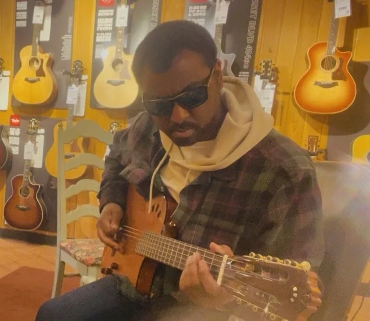 A man wearing sunglasses and a plaid jacket playing an acoustic guitar in a music store with other guitars on the wall.