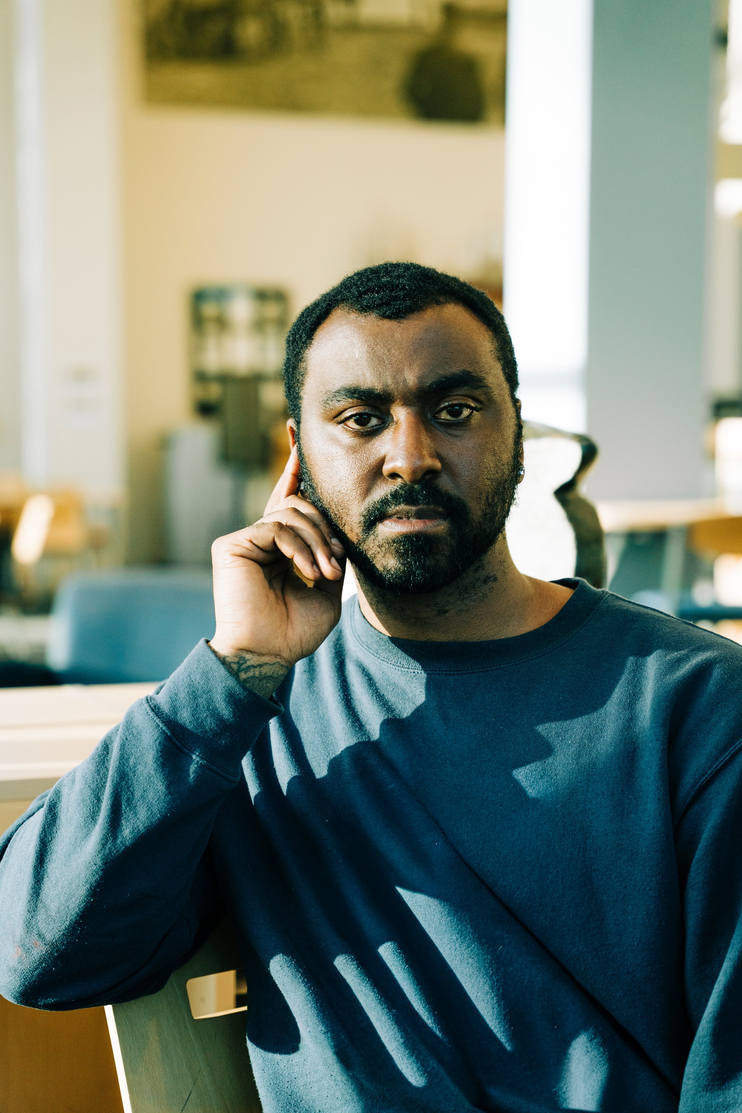 A man with dark skin, beard, and short curly hair is seated indoors, resting his face on his hand, wearing a dark long-sleeve shirt, with a focused or serious expression.