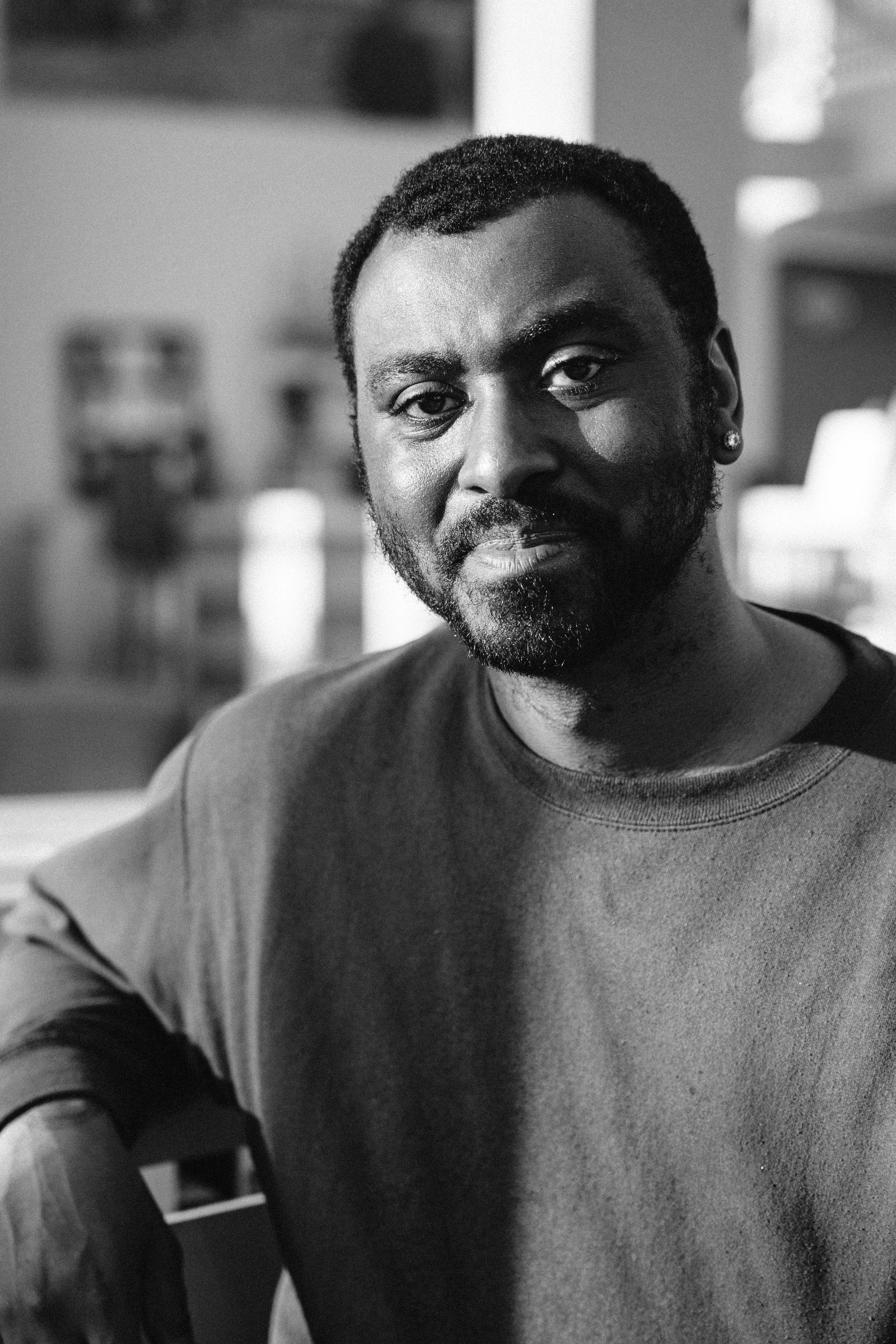 Black and white close-up portrait of a man with short curly hair, dark beard, wearing a casual dark t-shirt, sitting indoors with blurred background.