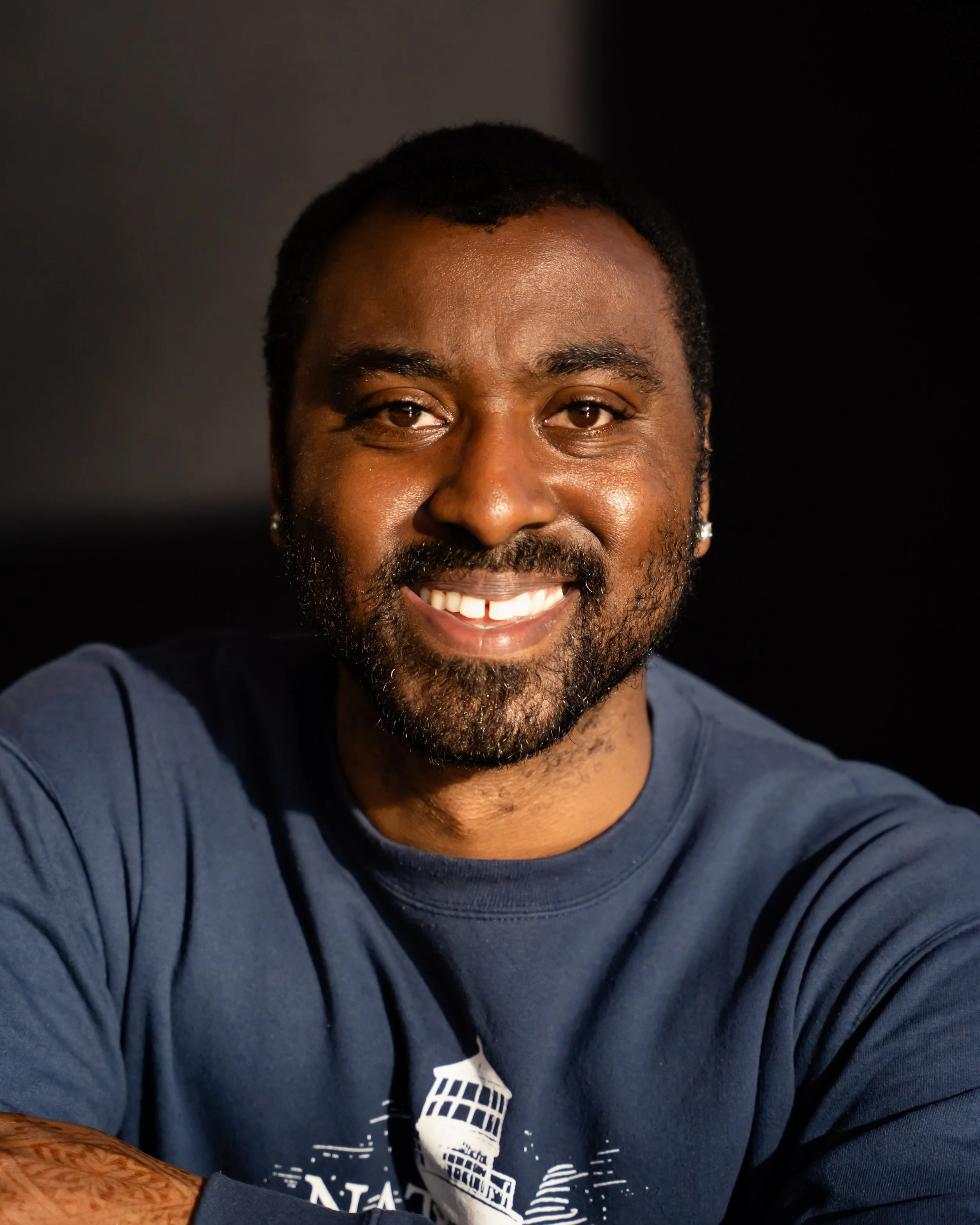 Close-up of a smiling Black man with short curly hair and a beard, wearing a navy blue shirt with a white graphic on it.