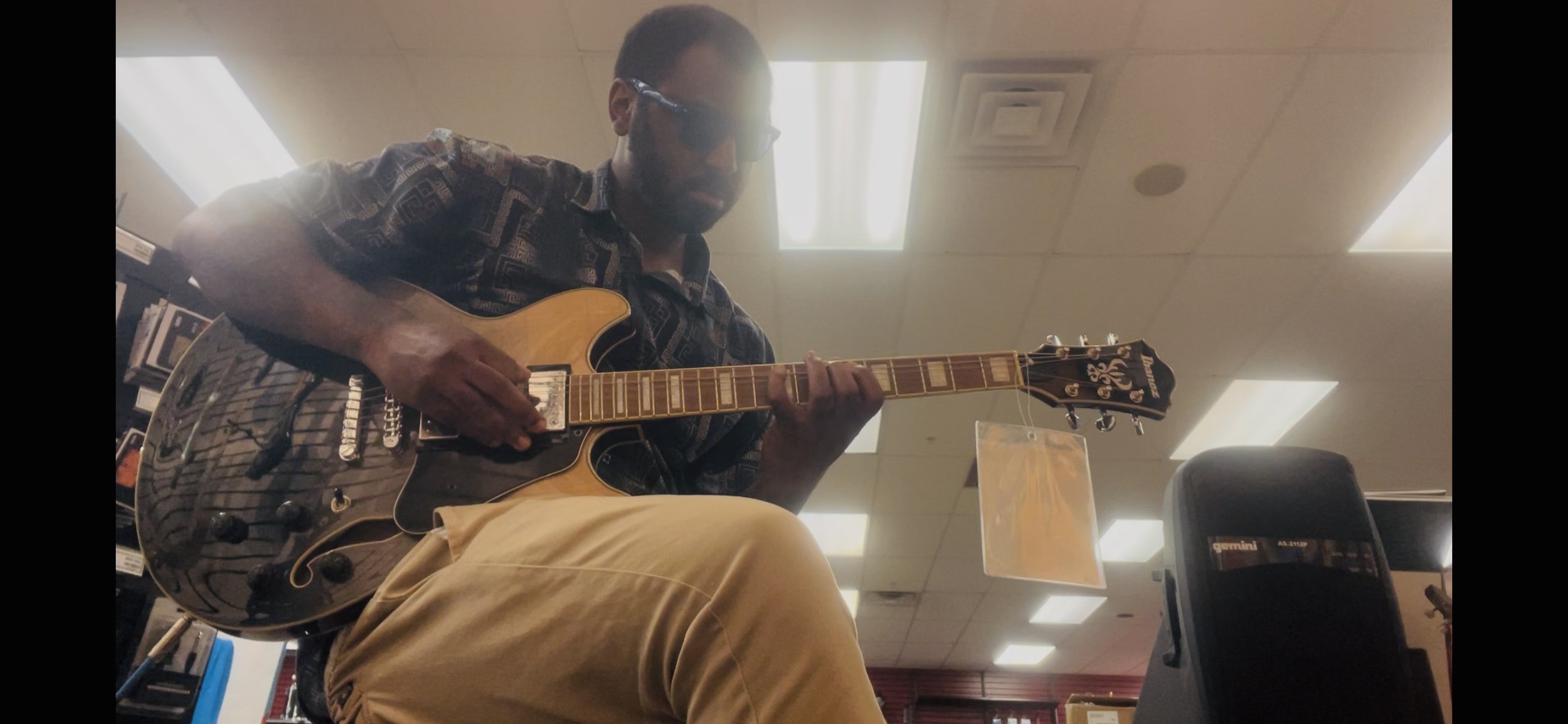 A man wearing sunglasses and a patterned shirt playing an electric guitar in an indoor setting, possibly a store or library.