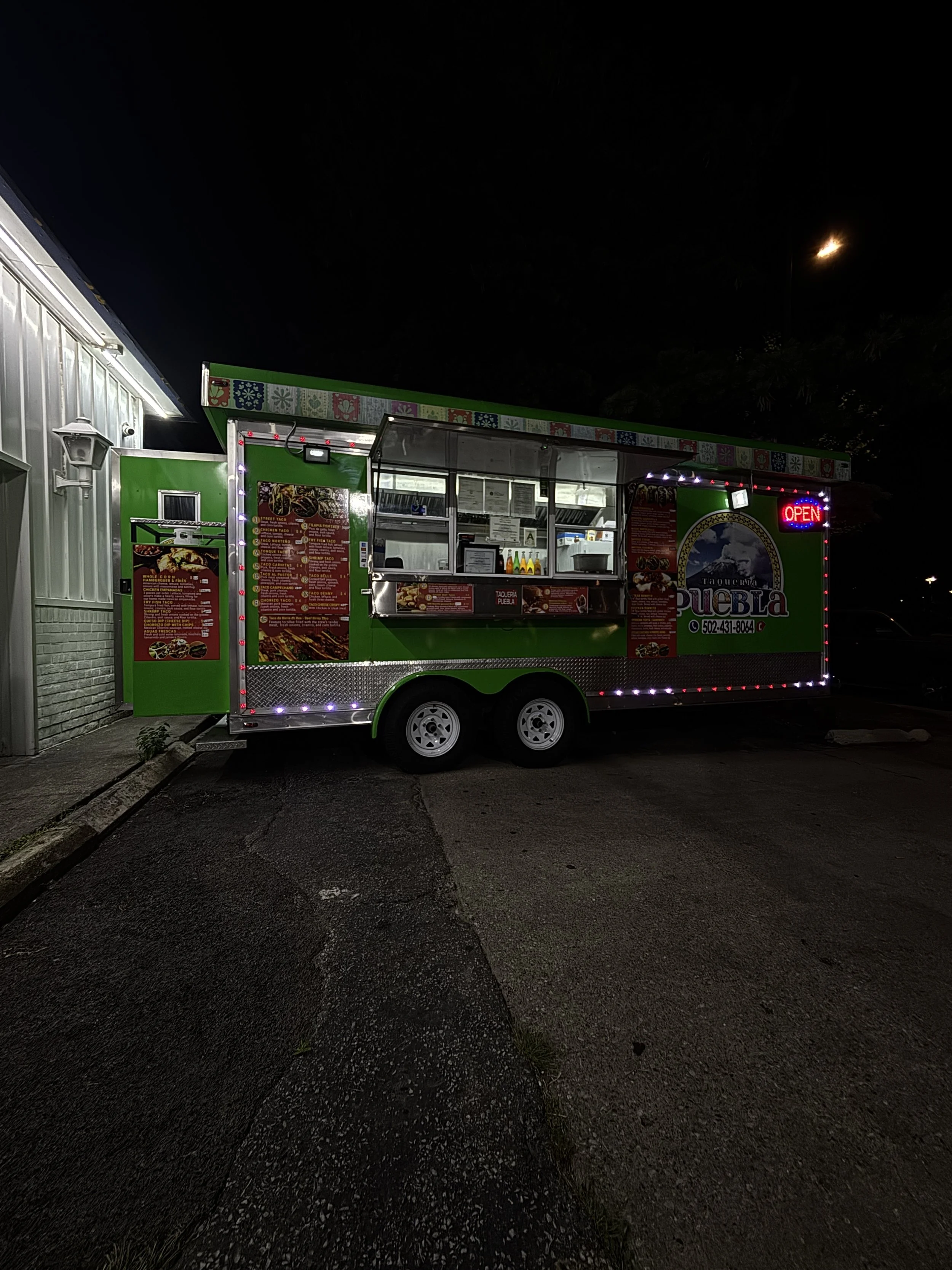 A brightly lit green food truck with a red 'OPEN' sign at night, parked on a dark street with a building and streetlight in the background.