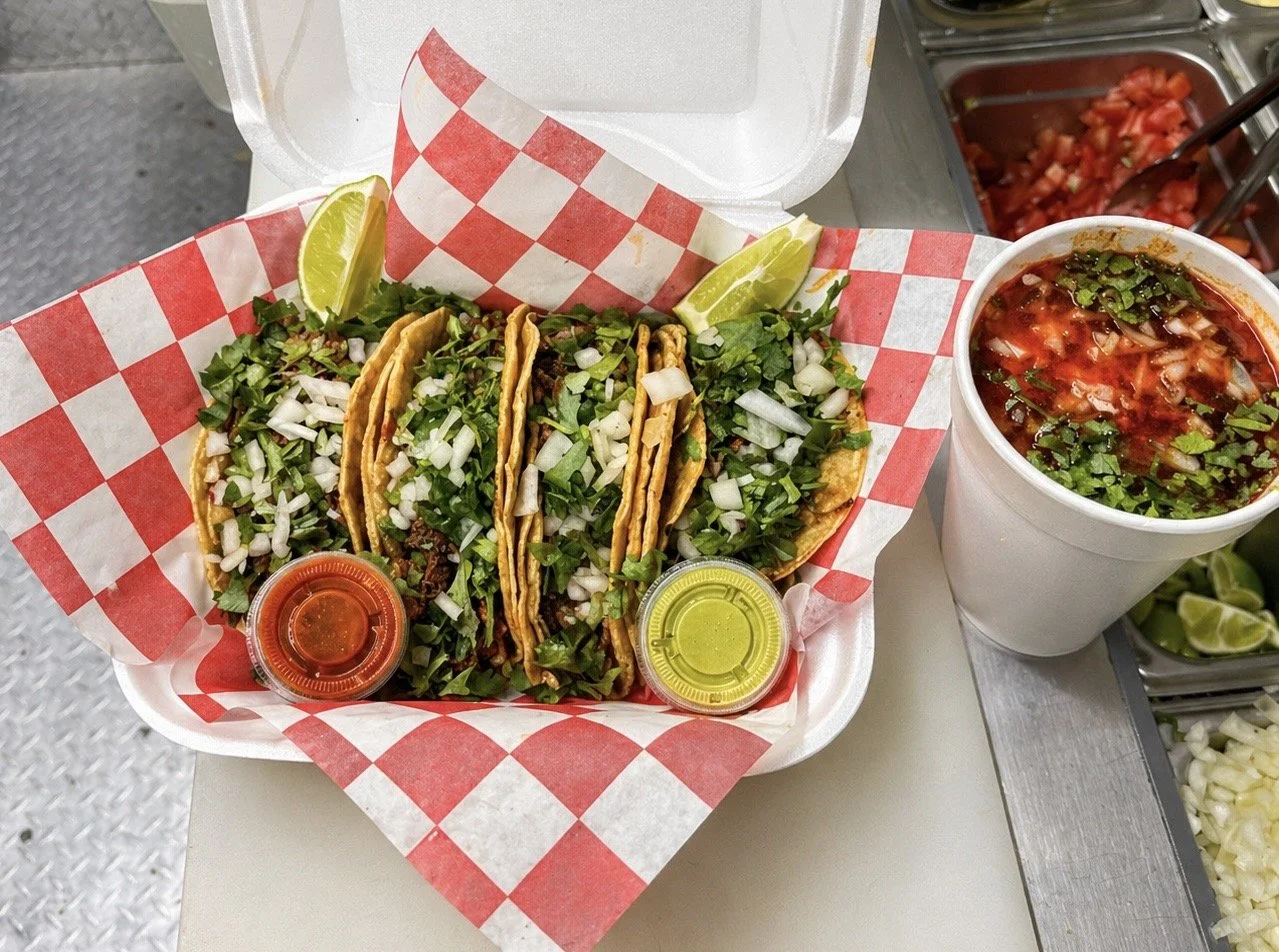 Four birria tacos filled with chopped onions, cilantro, and meat, served with lime wedges and hot sauce, alongside a cup of consume soup with cilantro on top.