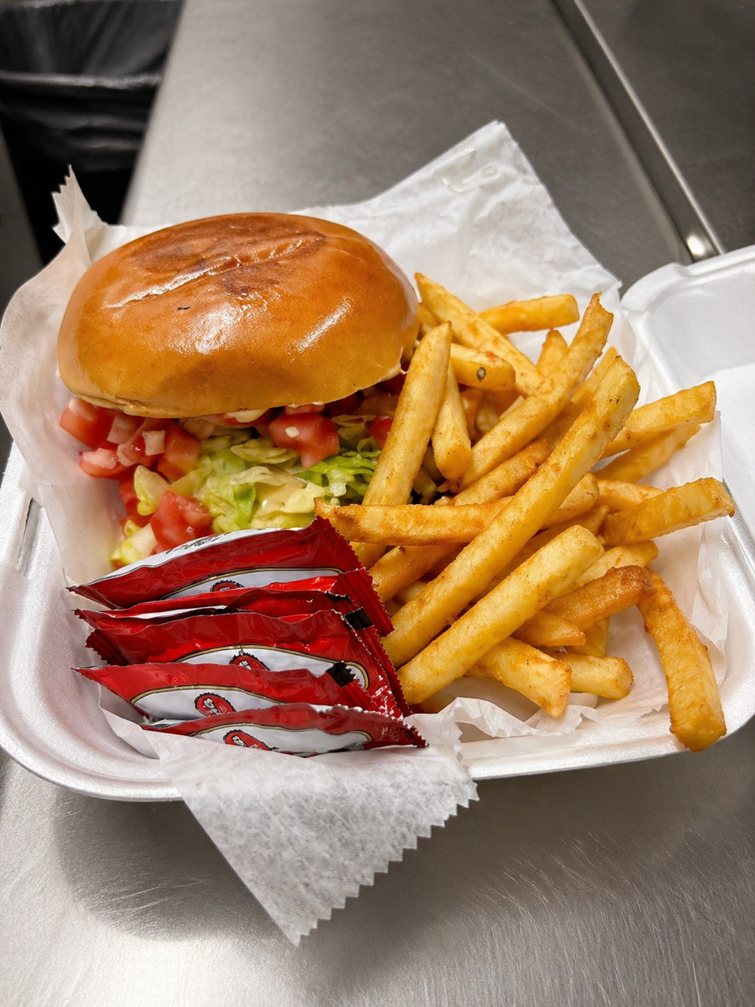 Foil container with a hamburger, French fries, ketchup packets, and chopped lettuce and tomatoes onions, and mayo inside the bun