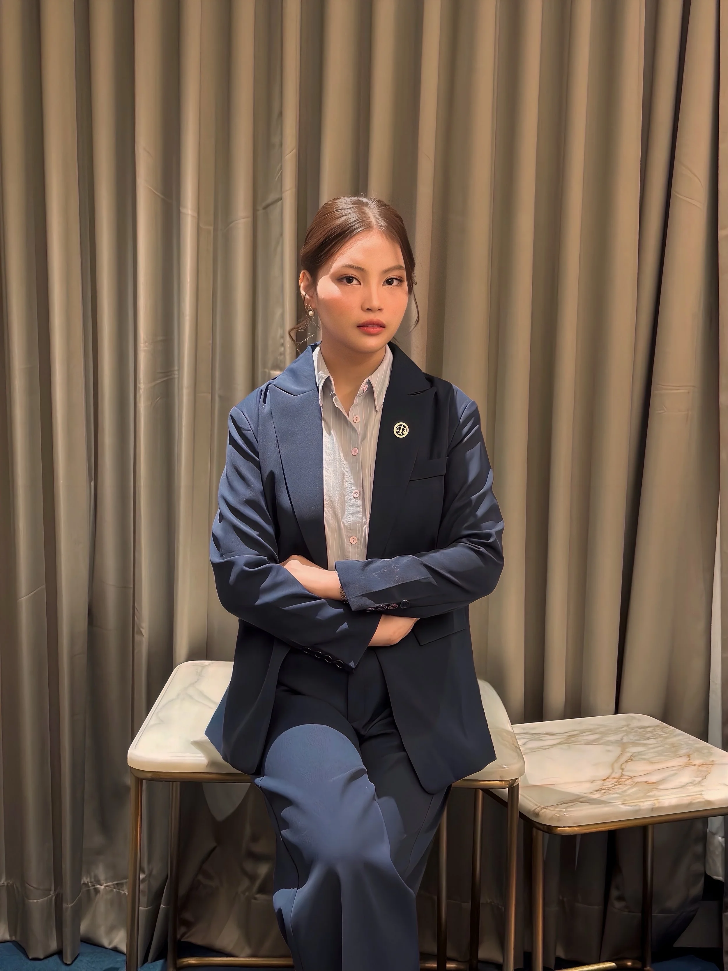 A woman with brown hair in a side part, wearing a white shirt and a dark blazer with a pin, sitting with her arms crossed on a marble table in front of beige curtains.