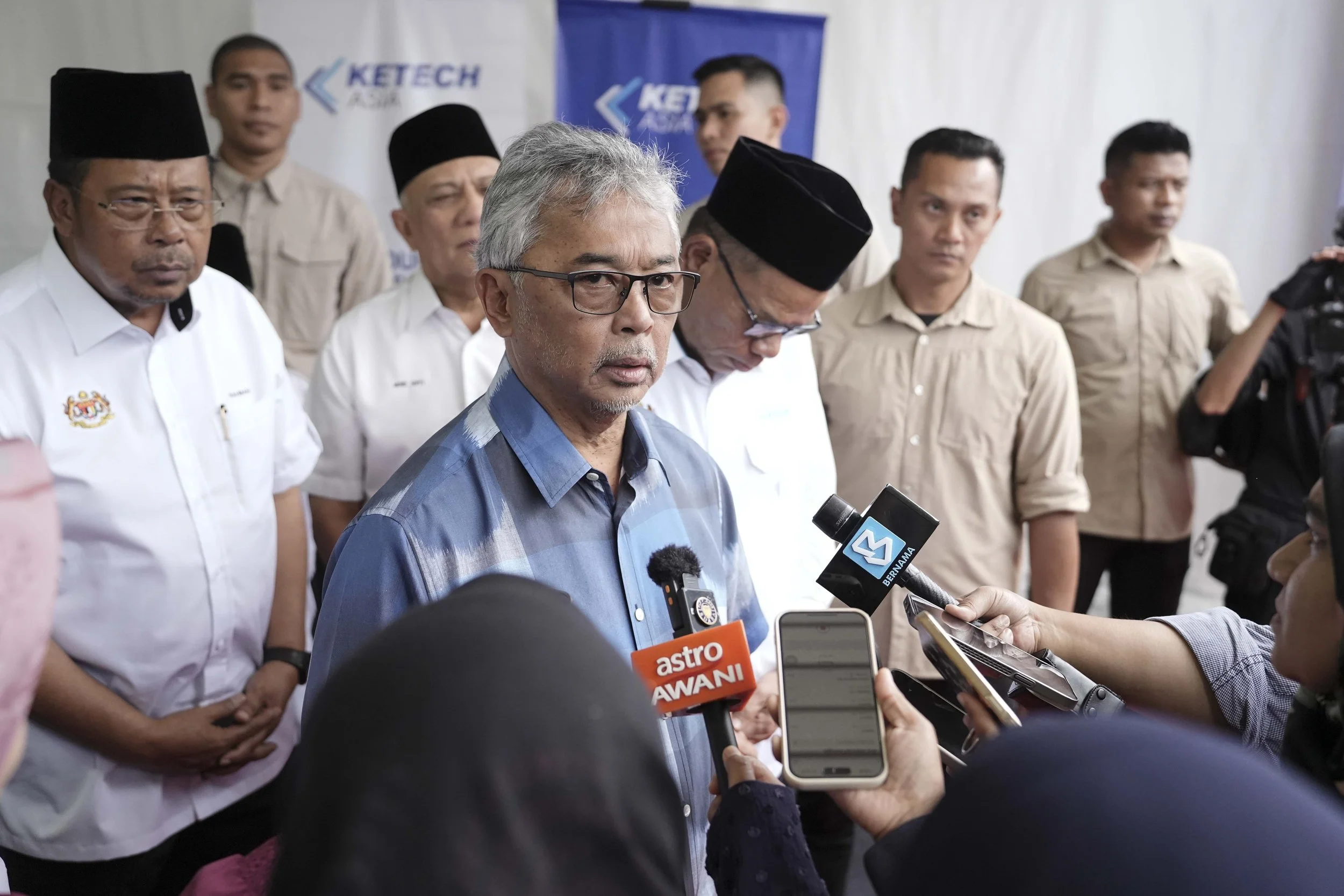 A group of men, some wearing traditional black hats, are standing behind a man with gray hair and glasses speaking to the media. The man is surrounded by microphones from various media outlets, including Astro Awani and Bernama, and appears to be giving a statement or interview.