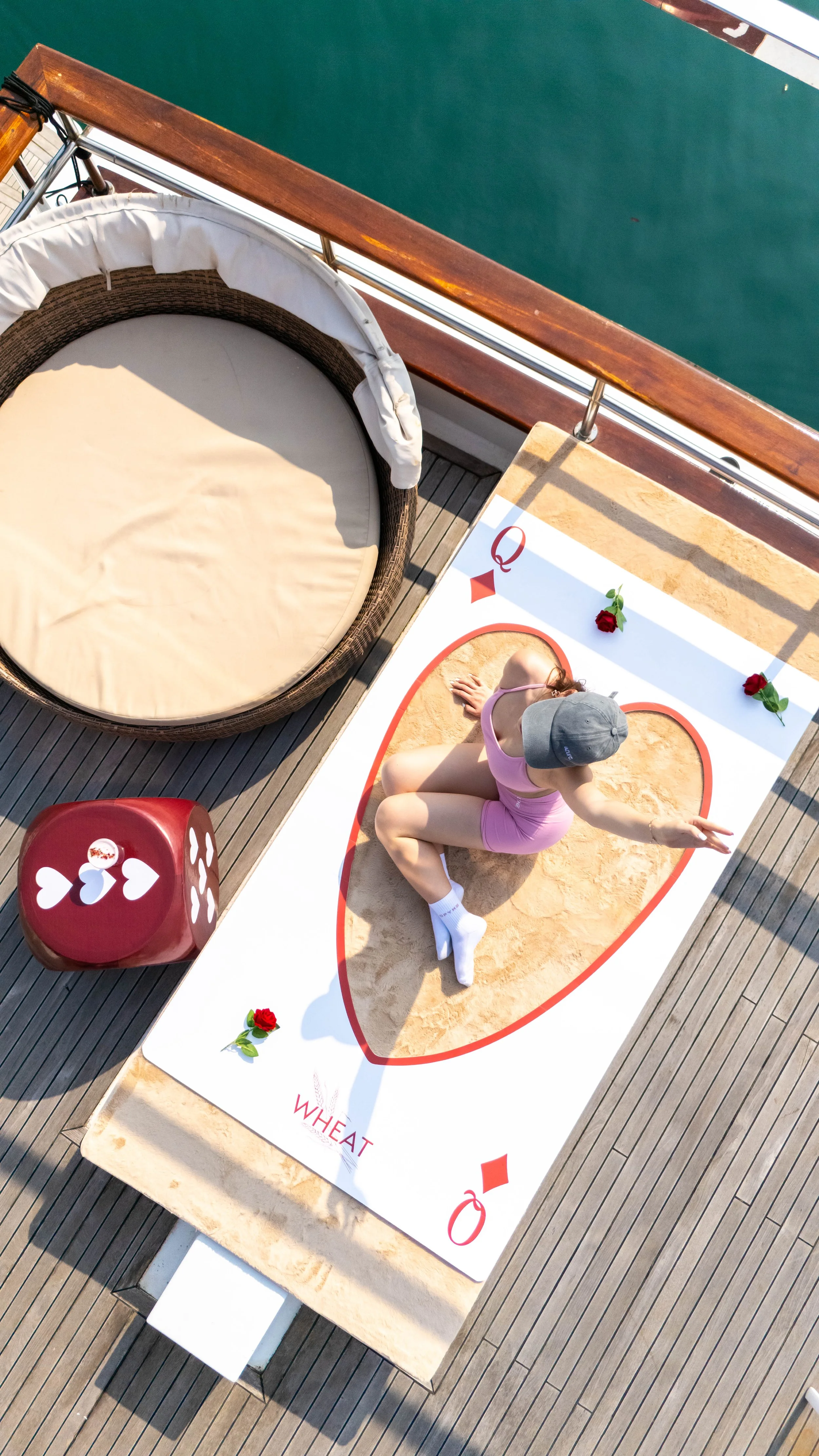 A woman in a pink athletic outfit and gray cap practicing yoga on a wheat-themed yoga mat on a deck, with a towel-covered round chair, giant red dice, and roses nearby, overlooking teal water.