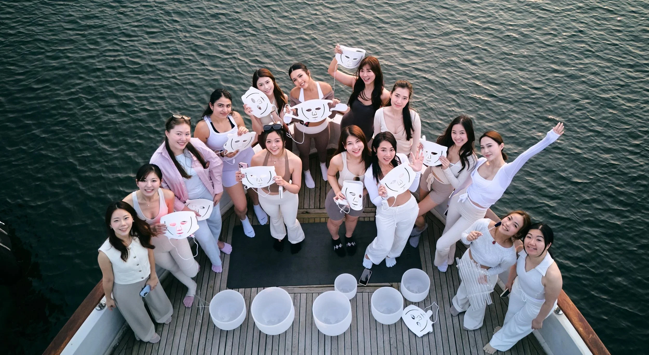 A group of women on a boat holding white masks with different facial expressions, smiling and waving at the camera.