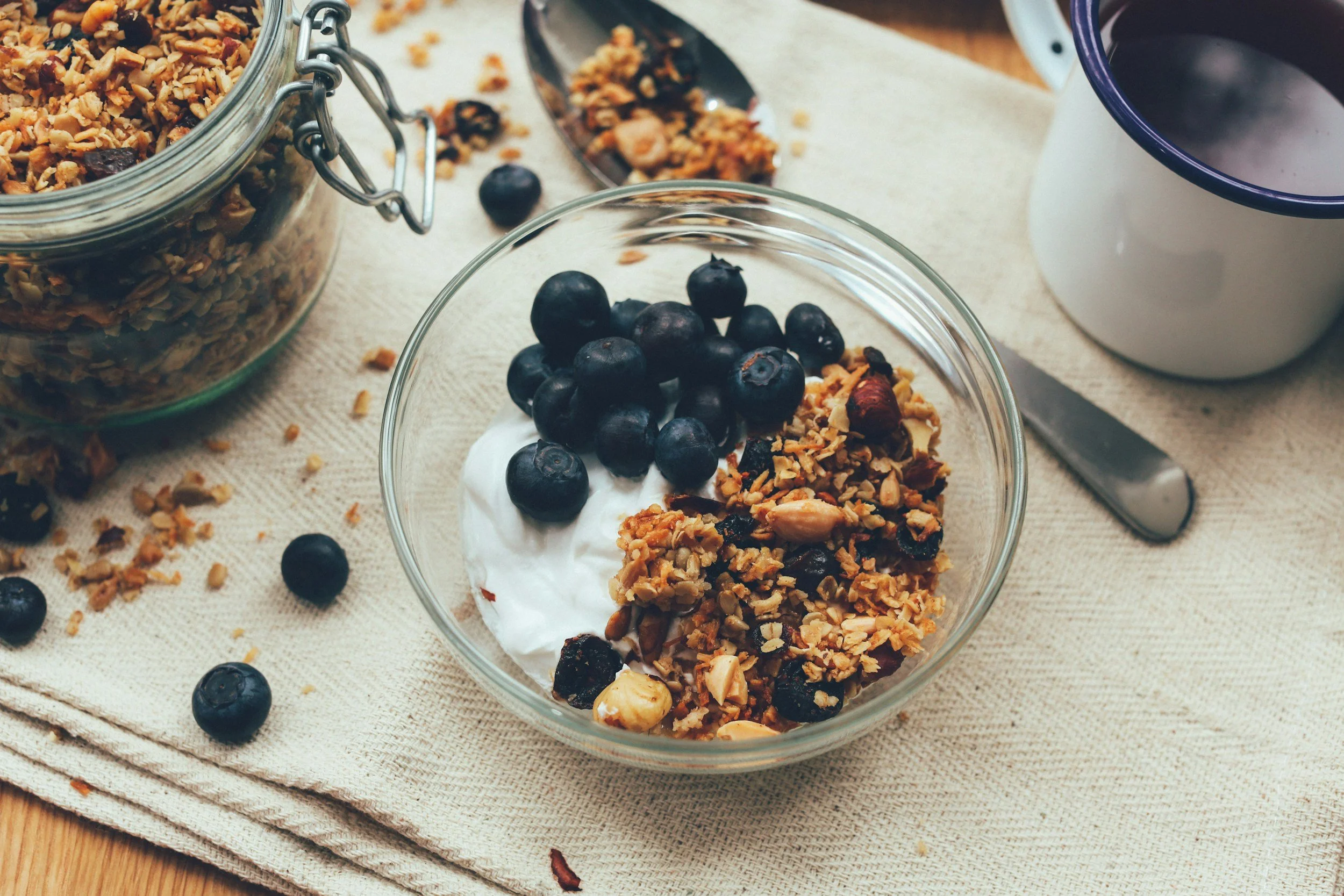 A glass bowl filled with yogurt topped with blueberries, granola, and nuts. Surrounding the bowl are scattered blueberries and granola on a light-colored cloth, with a jar of granola, a spoon, and a mug of coffee on a wooden surface.