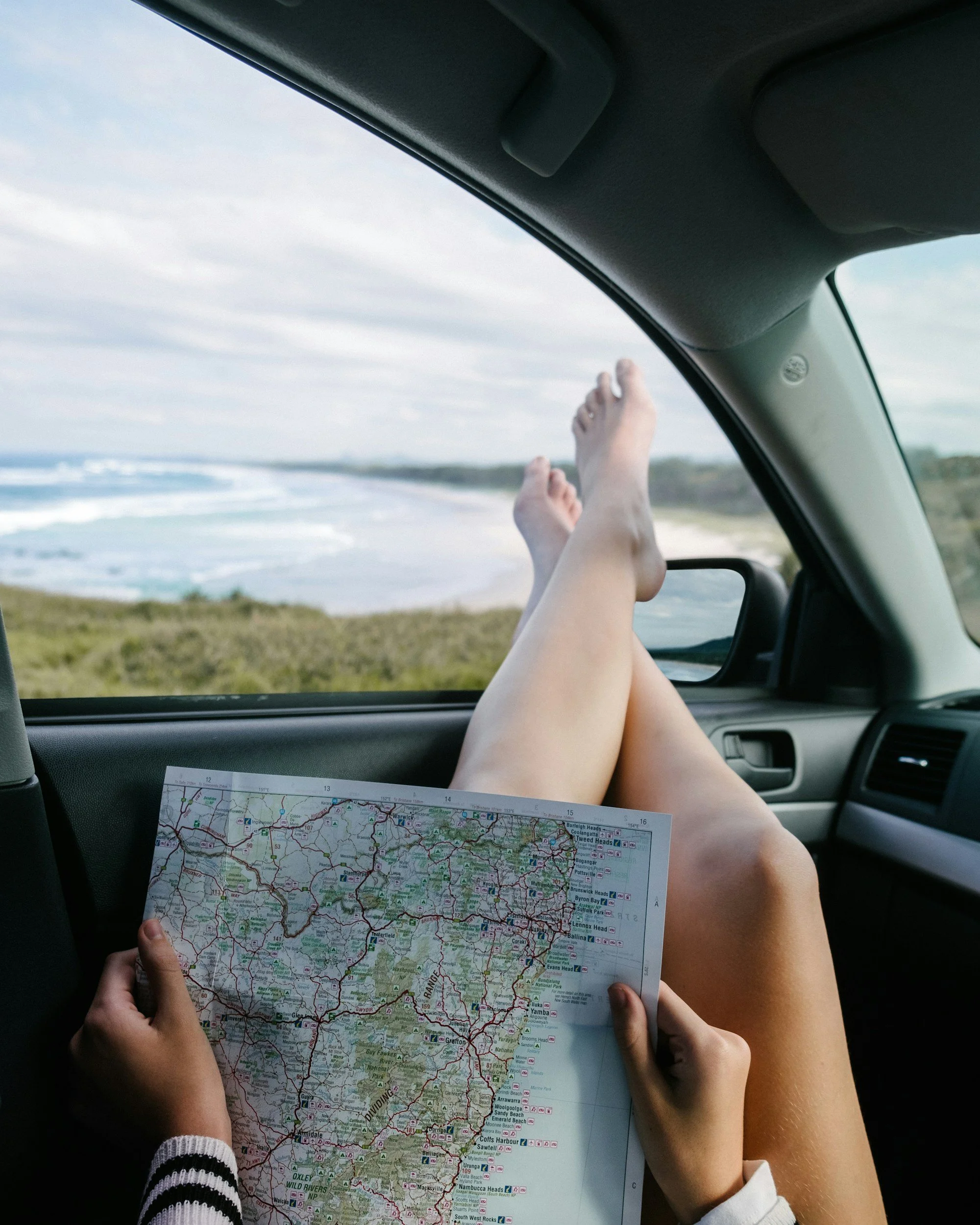 Person holding a map of New South Wales, Australia, while sitting inside a vehicle at the beach with legs extended outside the open window, with ocean and sky in the background.