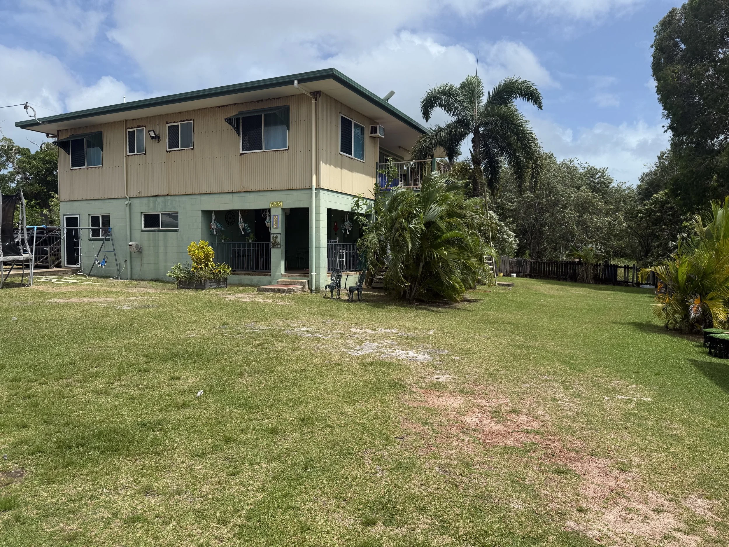 A two-story house painted in beige and light green, surrounded by tropical plants and trees, with a grassy yard and a trampoline on the left side. The house has multiple windows, a balcony, and an air conditioning unit on the upper floor.