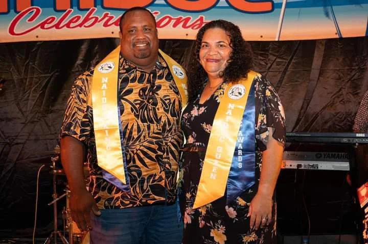 Two people, a man and a woman, stand together smiling, wearing awards sashes, at an event with a colorful backdrop. The man is wearing a black and gold shirt, and the woman is wearing a black dress with a floral pattern.
