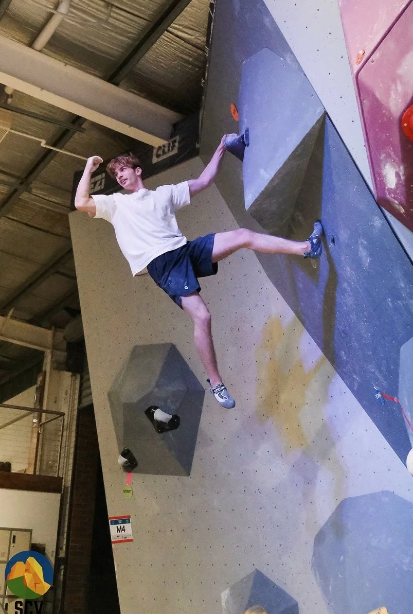 Young man in white t-shirt and navy shorts rock climbing on an indoor bouldering wall, flexing his arm and smiling.